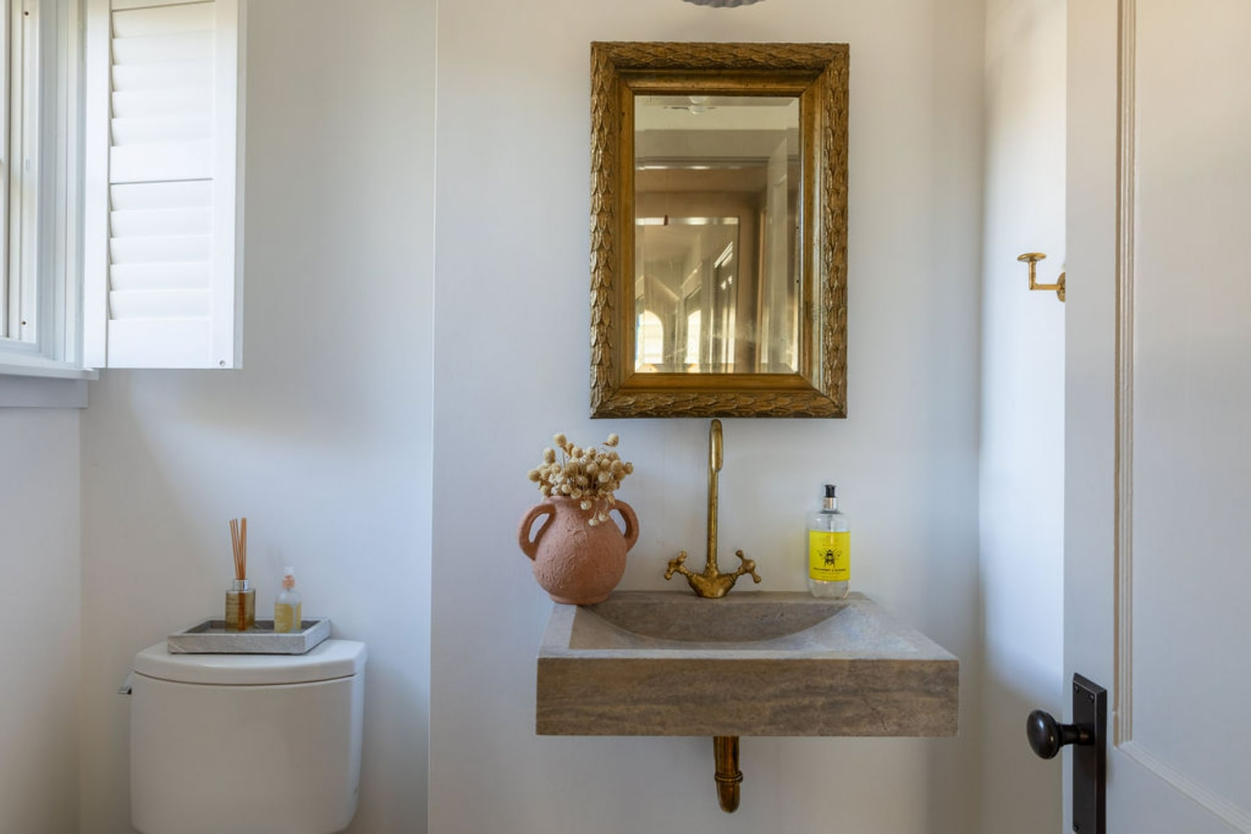 A bright powder room features a floating stone sink with a gold faucet, an ornate gold-framed mirror, and a toilet next to a window with white shutters.