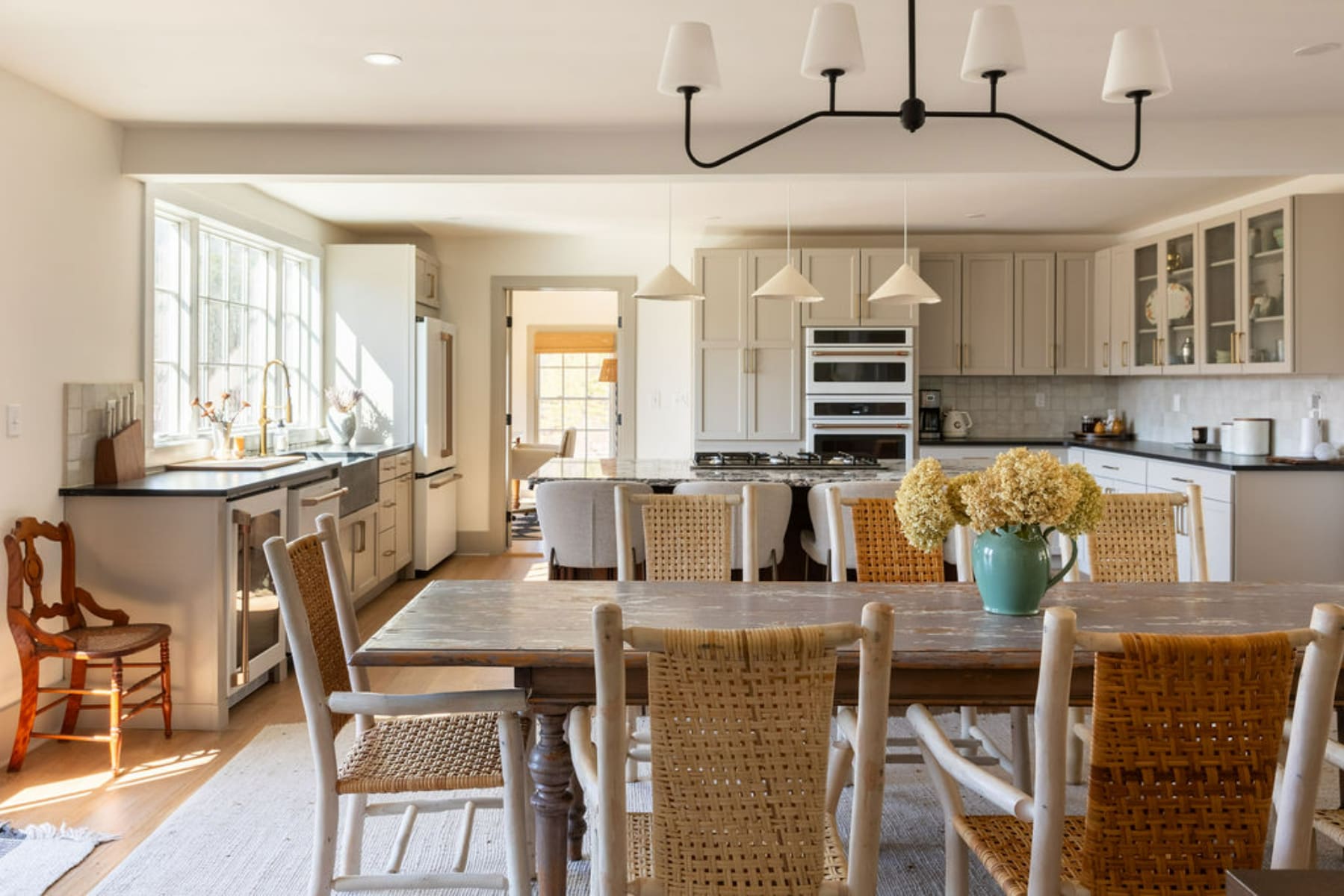 Open-concept kitchen and dining area featuring light gray cabinetry, a large dining table with mixed chairs, and a kitchen island with bar seating. A modern chandelier hangs over the dining table.