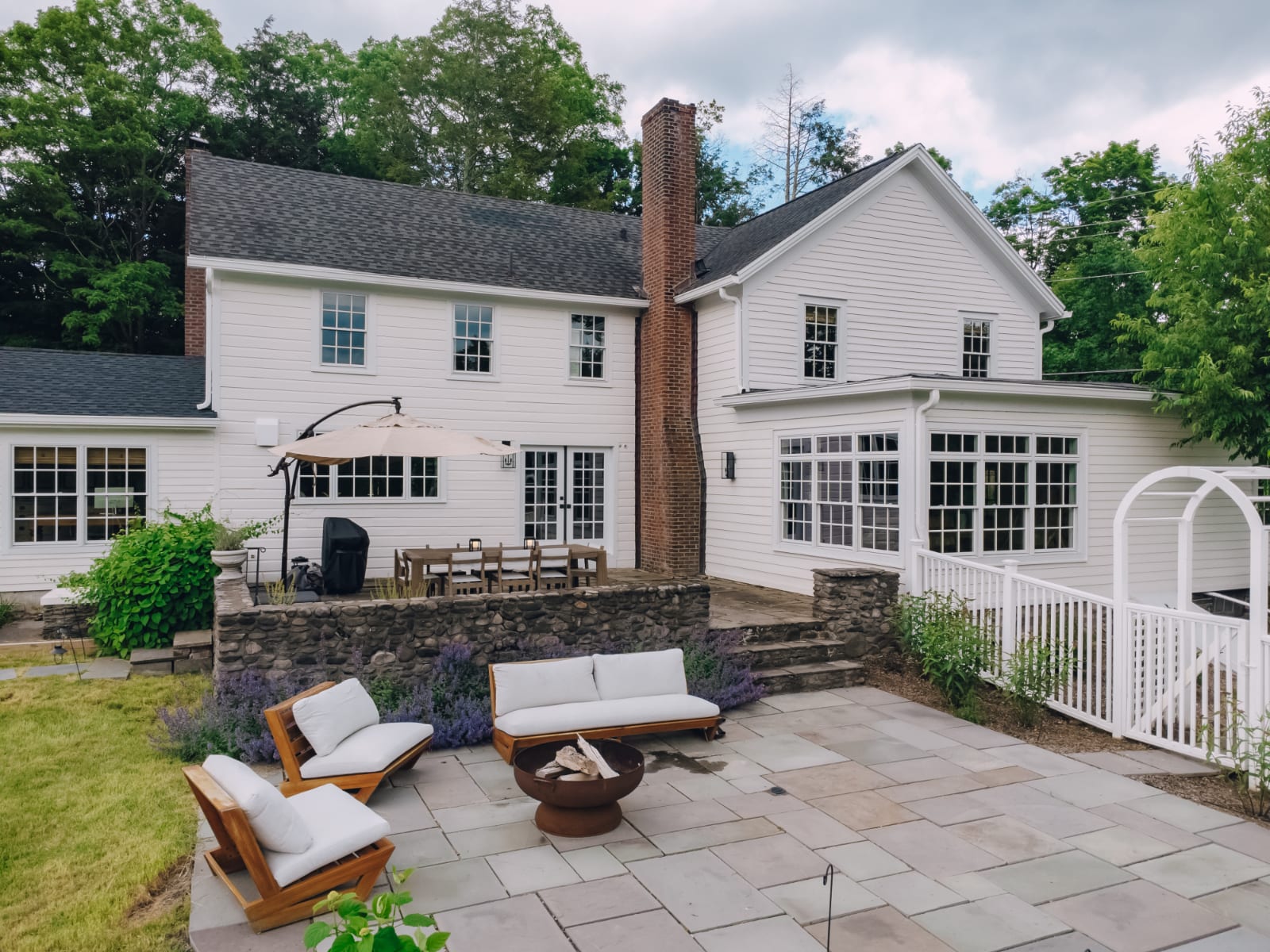 The back of a classic white farmhouse with multiple windows and a prominent stone chimney, overlooking a spacious stone patio. The patio features outdoor seating, a fire pit, and an outdoor dining area, surrounded by lush green lawn and mature trees.