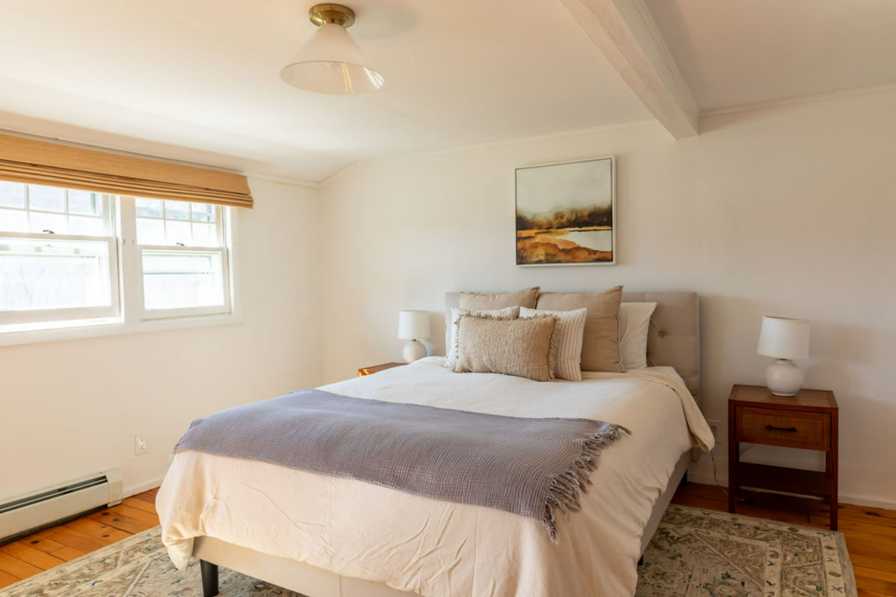 Inviting bedroom with a queen bed, flanked by wooden nightstands and lamps, set on a patterned area rug. A large window with a bamboo blind offers natural light, complementing the warm wooden floors.