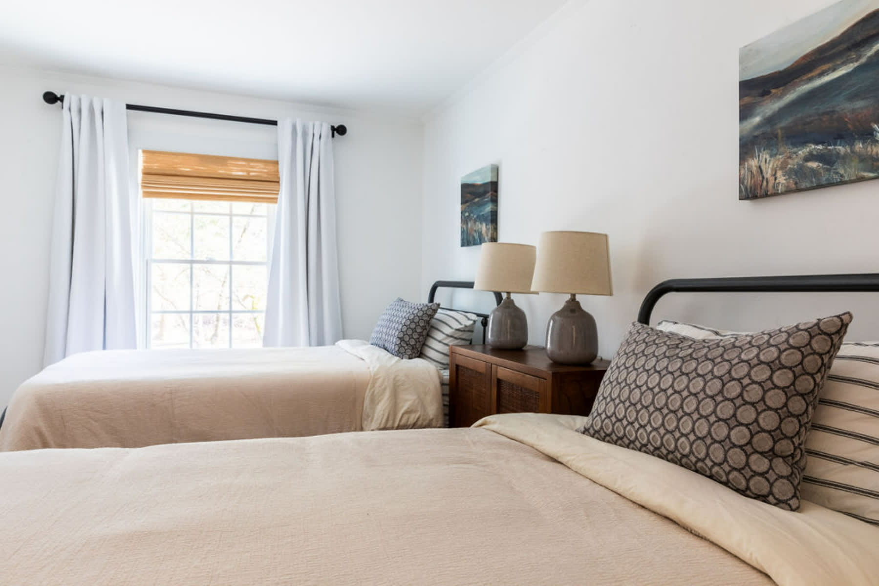 A bright bedroom featuring two black metal twin beds with neutral quilted bedding and patterned throw pillows. A window with a bamboo blind and white curtains provides natural light, flanked by a wooden nightstand with two lamps.