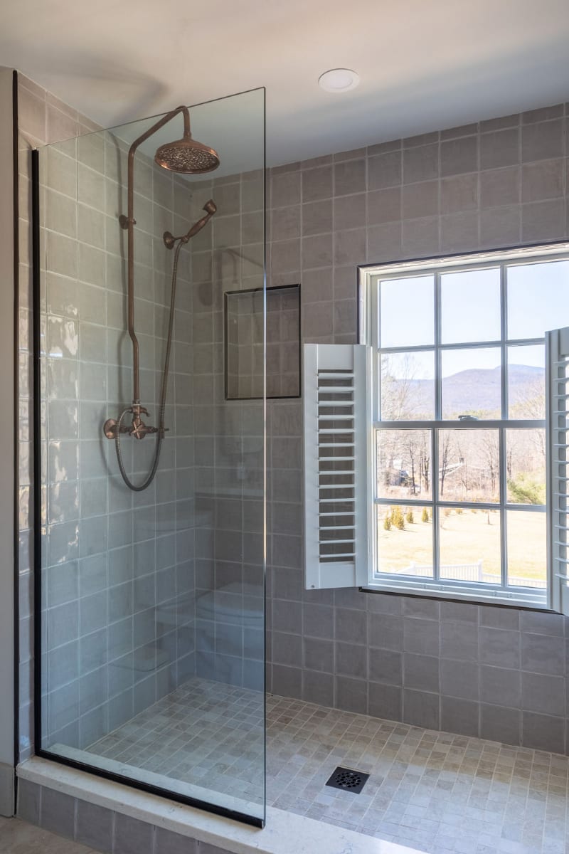 A modern walk-in shower with a clear glass enclosure, featuring bronze fixtures and light gray square tiles. A large window with white louvered shutters offers a scenic view of distant mountains and trees.