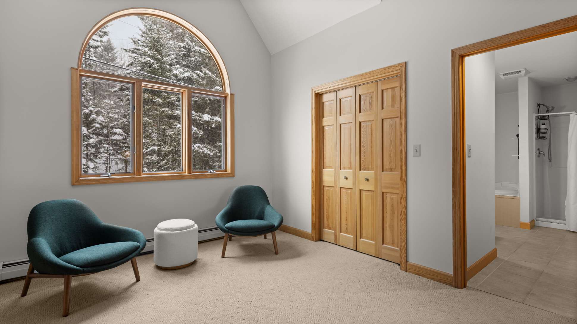 Corner view of the spacious bedroom highlighting the large arched window with wooden trim, offering stunning views of the snowy forest, alongside two teal armchairs and a white ottoman, with a closet door visible.