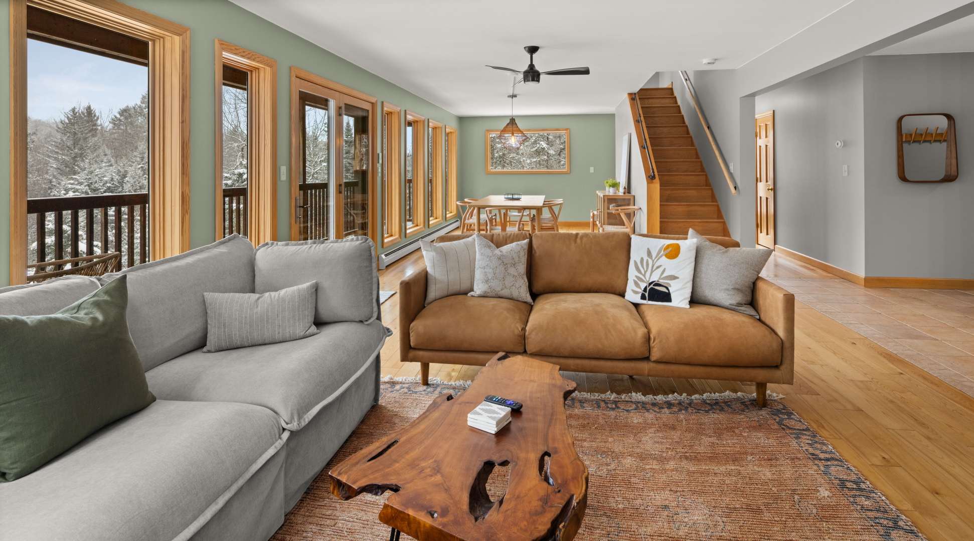 Expansive living area with light green walls, a brown leather sofa, a light gray sectional, and a unique live-edge coffee table, opening into a dining area with a wooden table and chairs, all with views of a snowy forest through numerous windows.