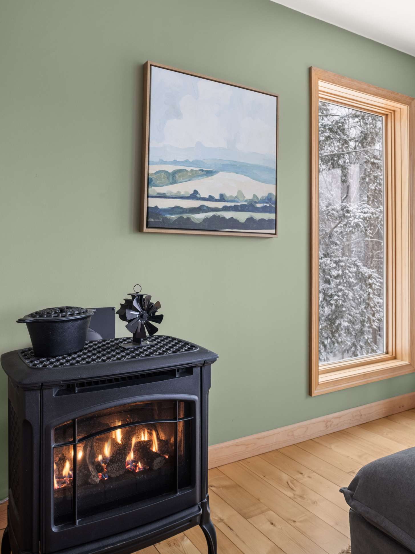A cozy corner of the living space features a black wood-burning stove with a small fan on top, set against a sage green wall. A large window with a natural wood frame offers a picturesque view of a snow-covered forest.