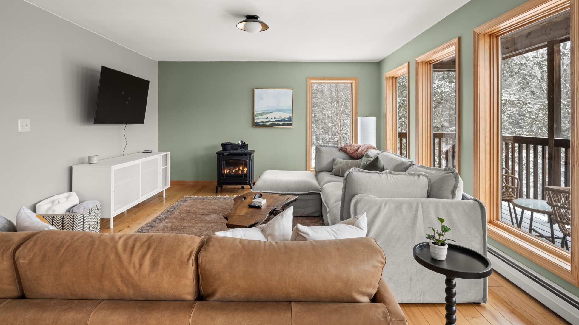 Cozy living room with light green walls, a large L-shaped sectional sofa, a brown leather sofa, and a rustic live-edge coffee table, featuring a modern electric fireplace and large windows overlooking a snowy landscape.