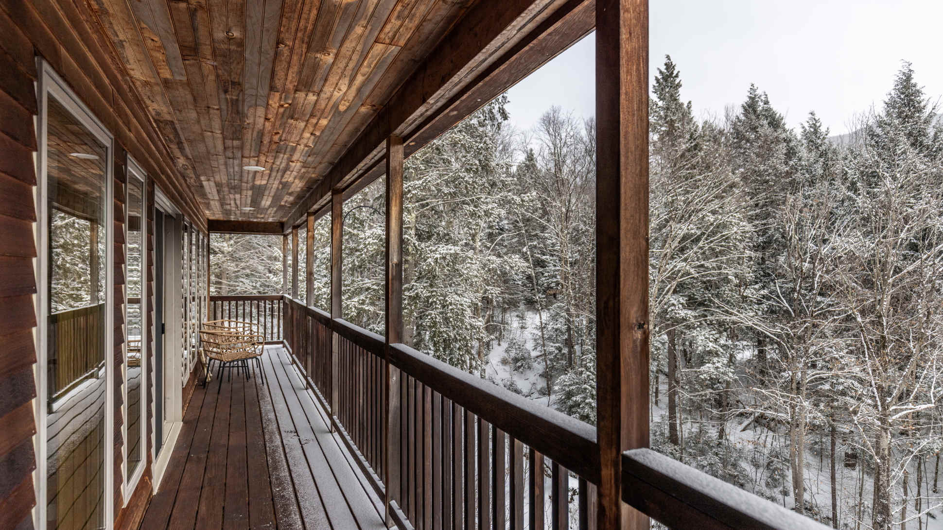 A rustic wooden balcony extends along the side of the house, offering expansive views of a snow-covered evergreen forest. The ceiling and railings are made of natural wood, creating a warm and inviting outdoor space.