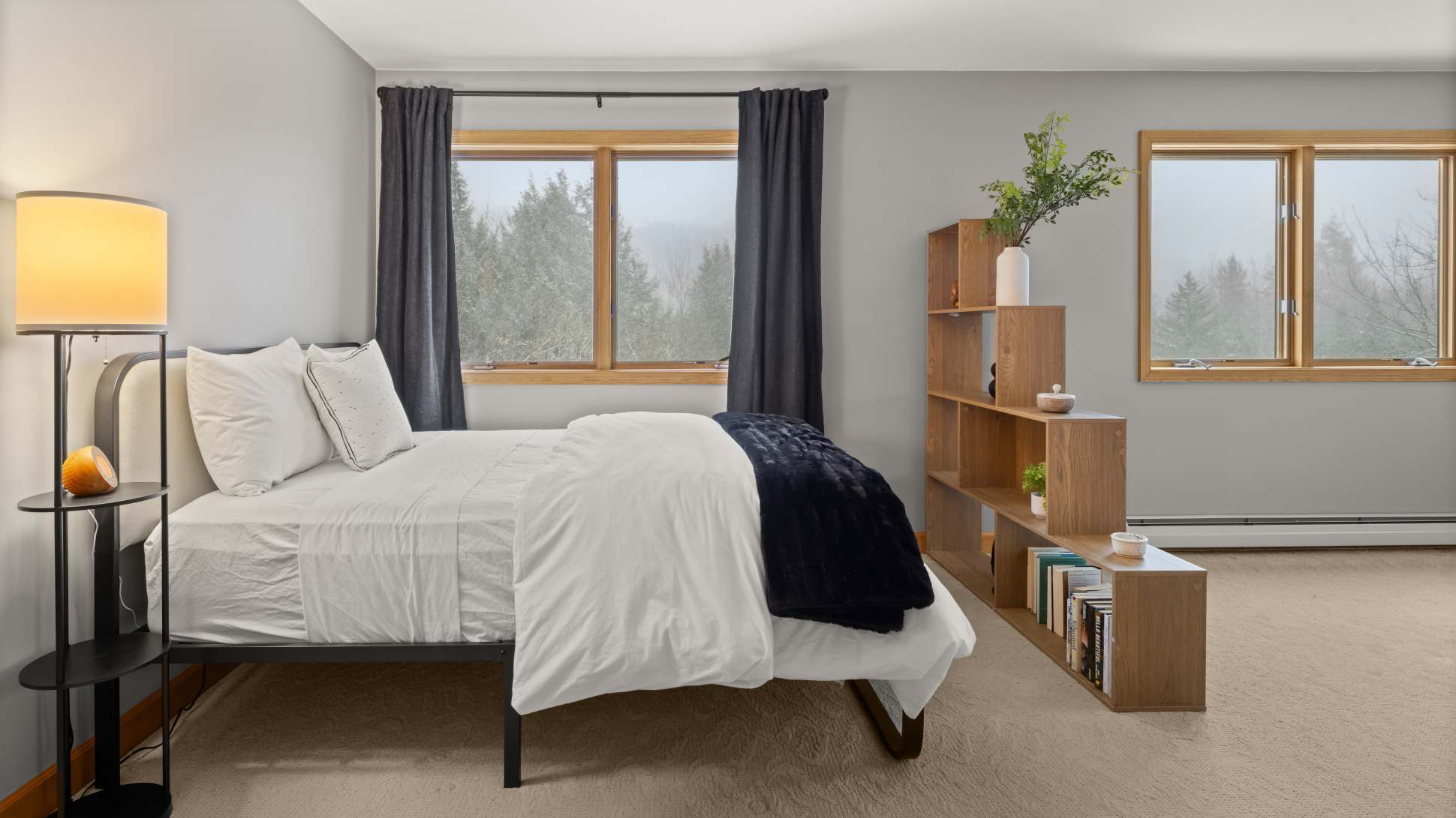 A bedroom with light grey walls and carpet, featuring a metal-framed bed with white bedding and a dark blue throw. A tall floor lamp stands beside the bed, and a wooden shelving unit with decor is visible.