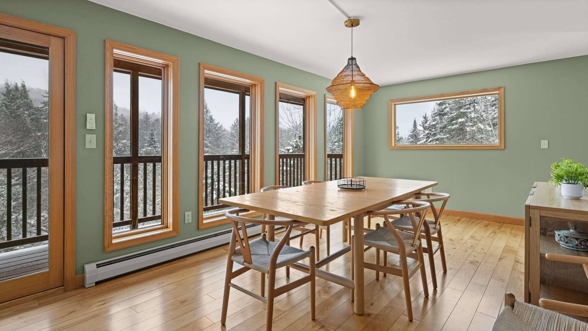Bright dining room with light green walls and hardwood floors, featuring a long wooden dining table surrounded by modern wooden chairs, illuminated by a woven pendant light, with multiple windows and a door leading to a balcony overlooking a snowy forest.