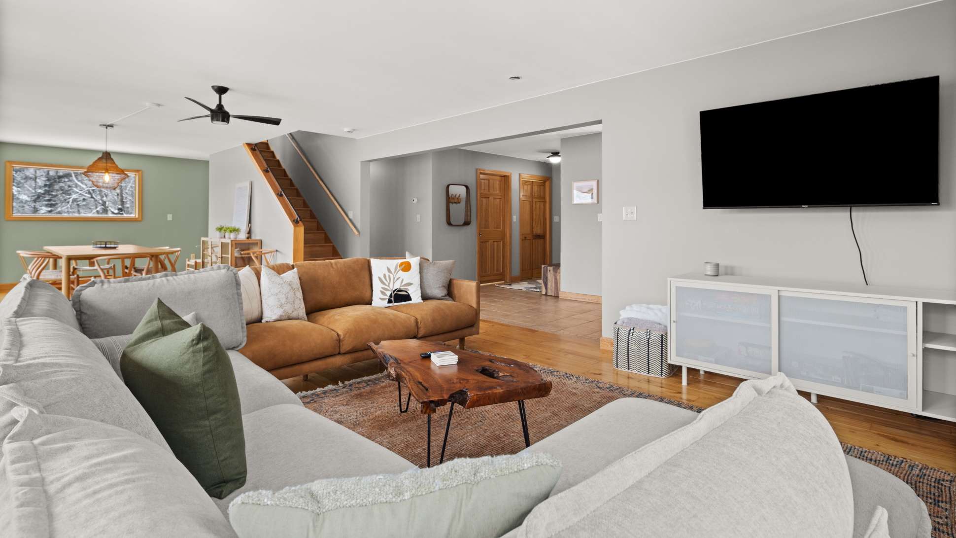 A spacious living room features a large L-shaped sectional sofa in light gray and a brown leather sofa, centered around a rustic live-edge wood coffee table on a patterned rug. A wall-mounted TV is visible, and a staircase leads to an upper level.