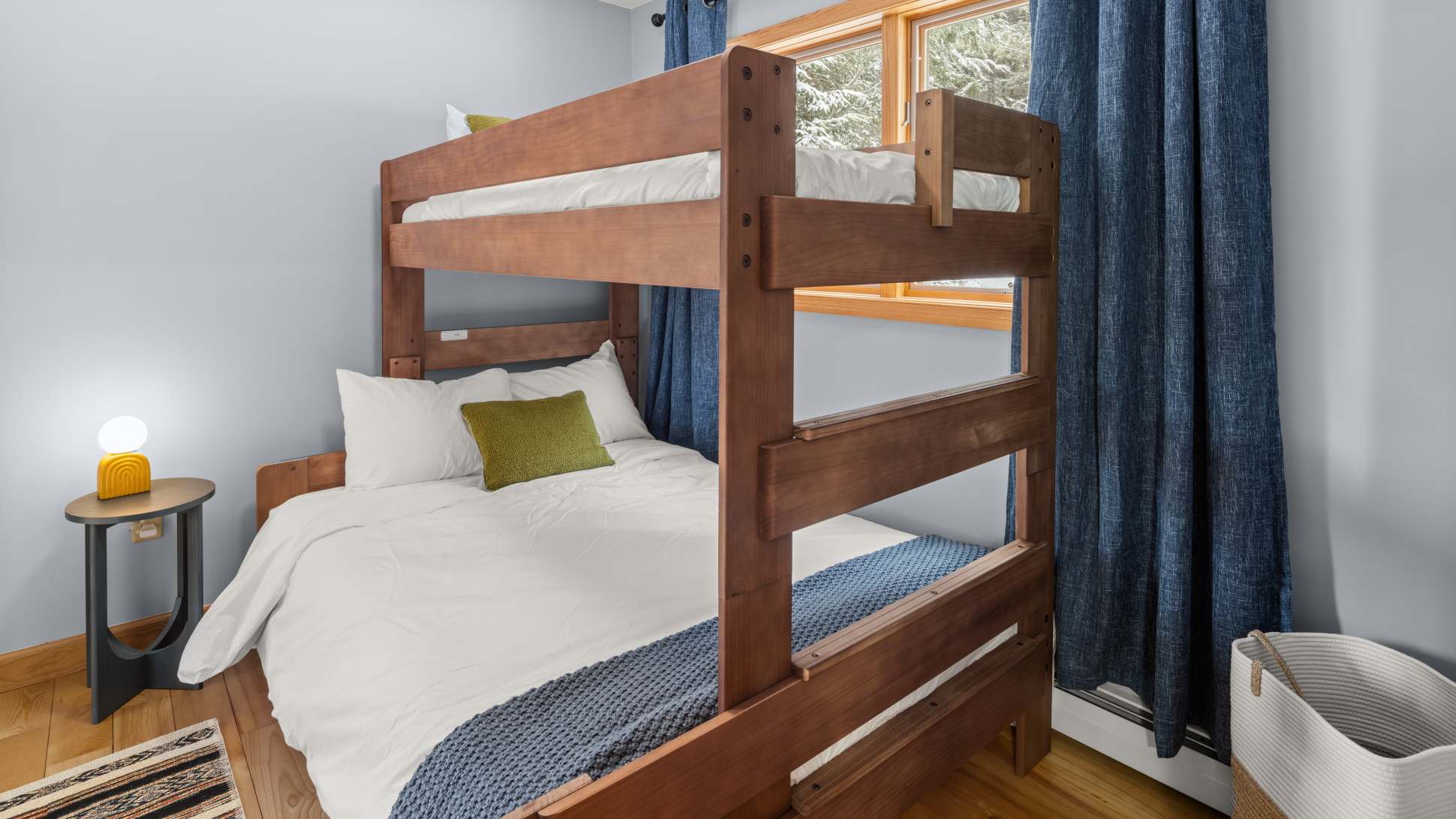 Detailed shot of the wooden bunk bed in a light blue bedroom, showing the full-size bottom bunk with white bedding and a green accent pillow, and a twin top bunk, with a window revealing a snowy forest outside.