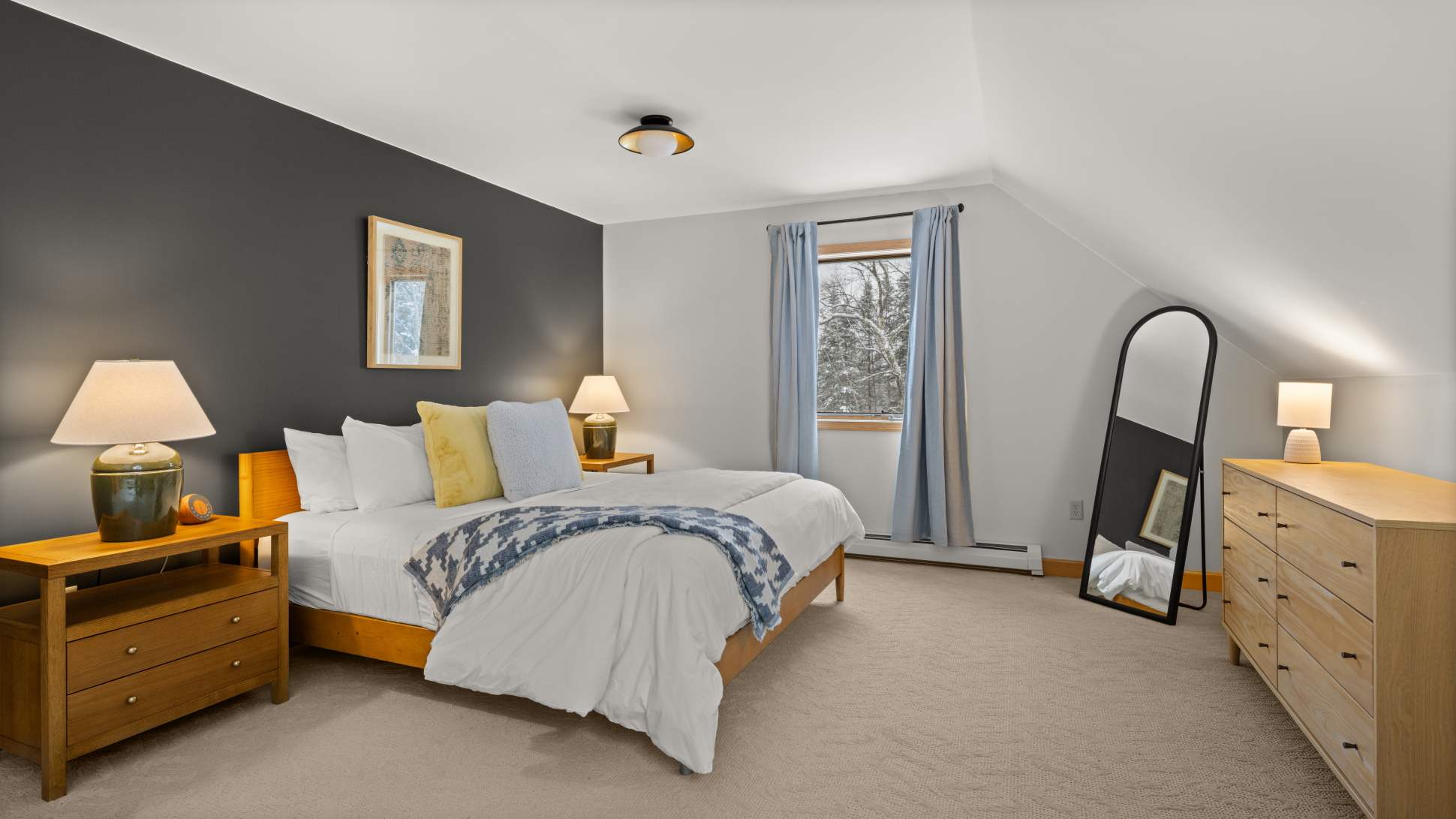 A spacious bedroom featuring a queen bed with a dark accent wall, flanked by wooden nightstands and lamps. A large mirror leans against the wall next to a light wood dresser, and a window with blue curtains offers a view of snowy trees.