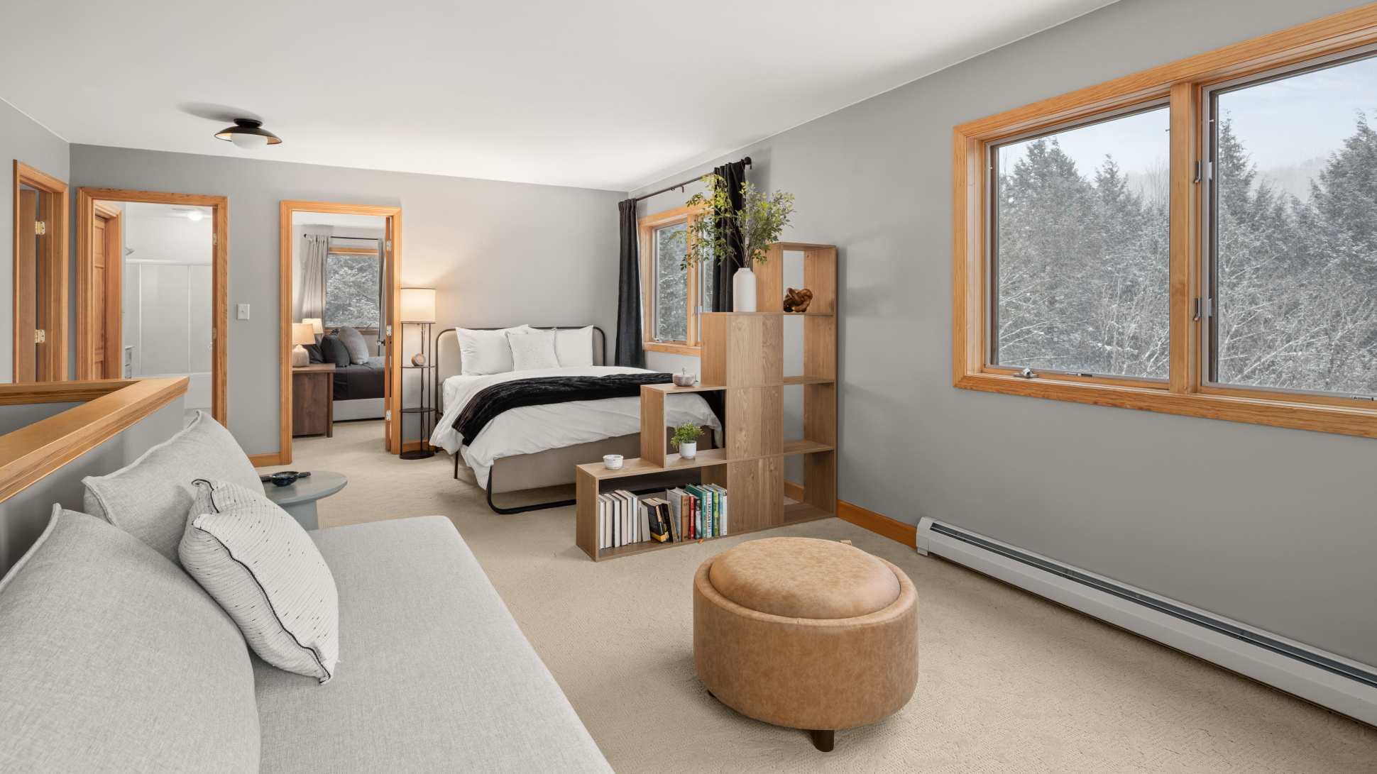 A spacious bedroom with light grey walls and carpeted flooring, featuring a bed with a dark metal frame, a light grey sofa, a round leather ottoman, and large windows offering a snowy forest view.