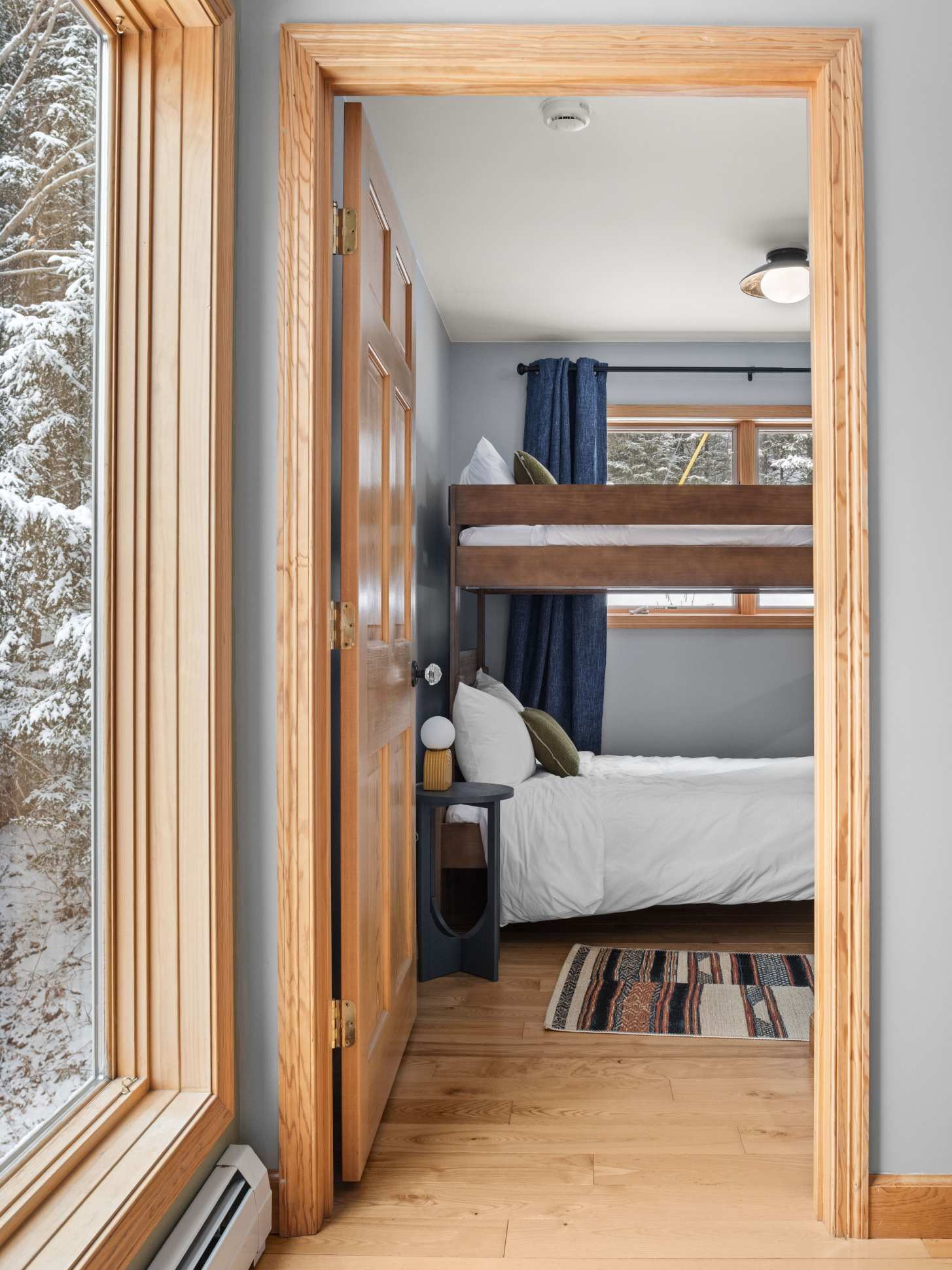 View into a cozy bedroom with light blue walls and hardwood floors, featuring a wooden bunk bed with a full-size bottom bunk and a twin top bunk, and a window offering a glimpse of the snowy outdoors.