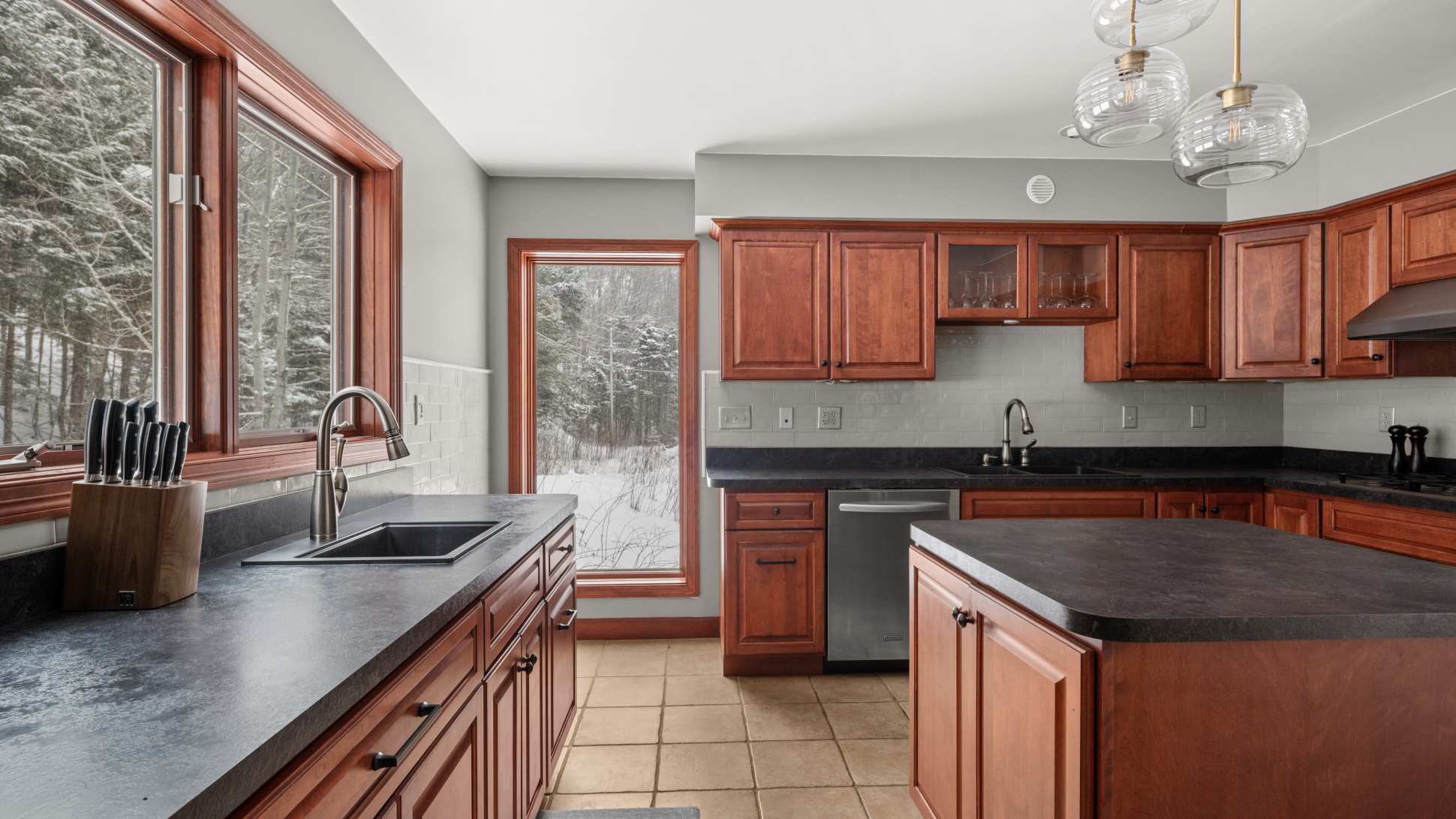 View of the kitchen featuring dark countertops, cherry wood cabinets, and a light gray subway tile backsplash, with a double sink under a large window overlooking a snowy forest, and a kitchen island in the foreground.