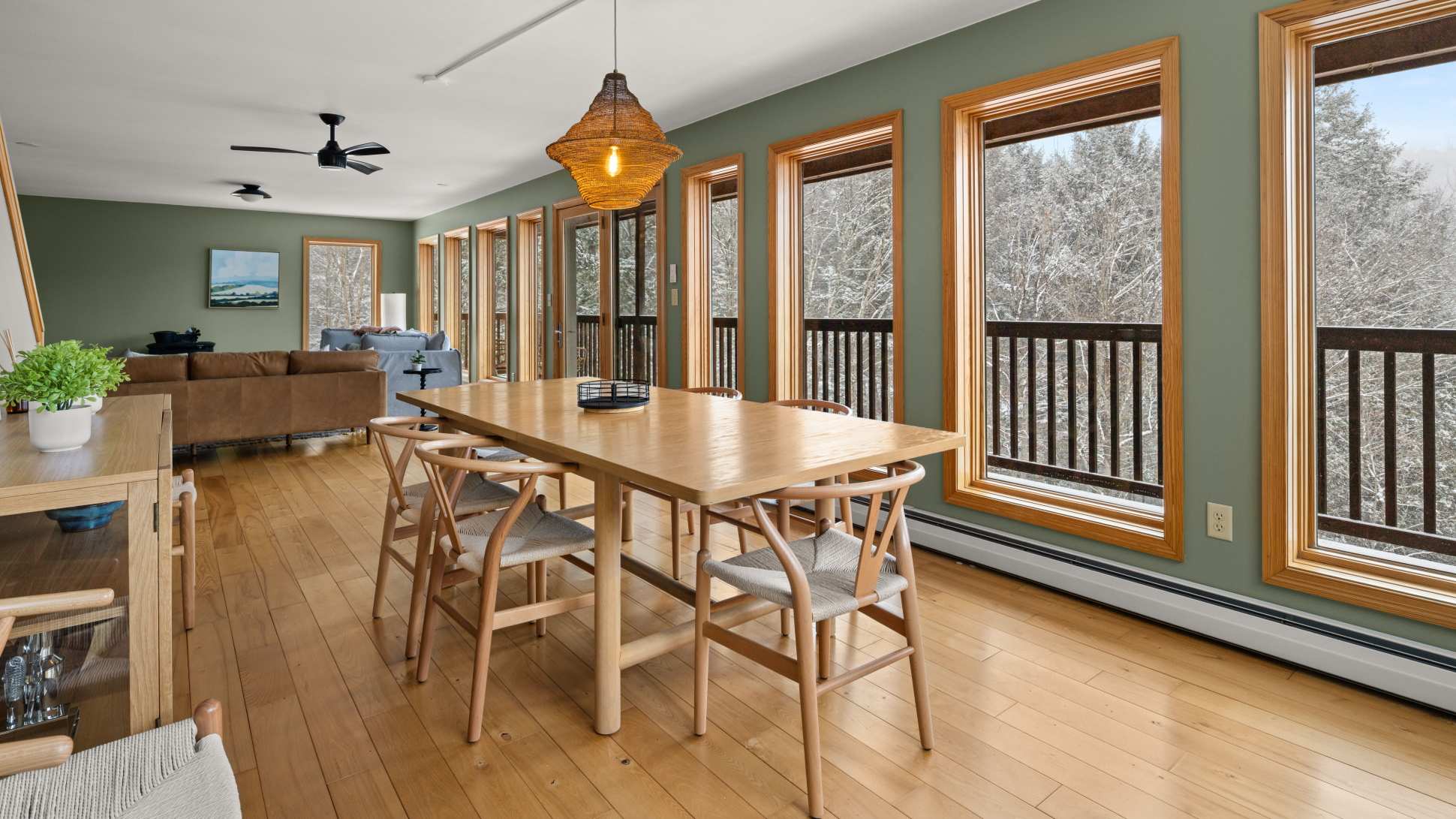 A bright dining room features a large light wood dining table with six matching chairs, set against a sage green wall with multiple windows offering snowy forest views. A woven pendant light hangs above the table, and hardwood floors extend throughout the space.