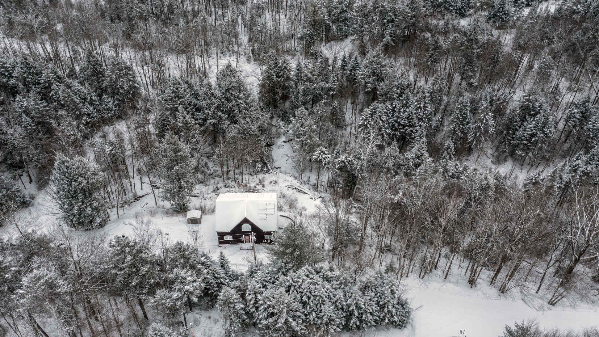 An aerial view captures a dark-colored house with a snow-covered roof nestled amidst a dense, snow-dusted forest, showcasing its secluded winter setting.