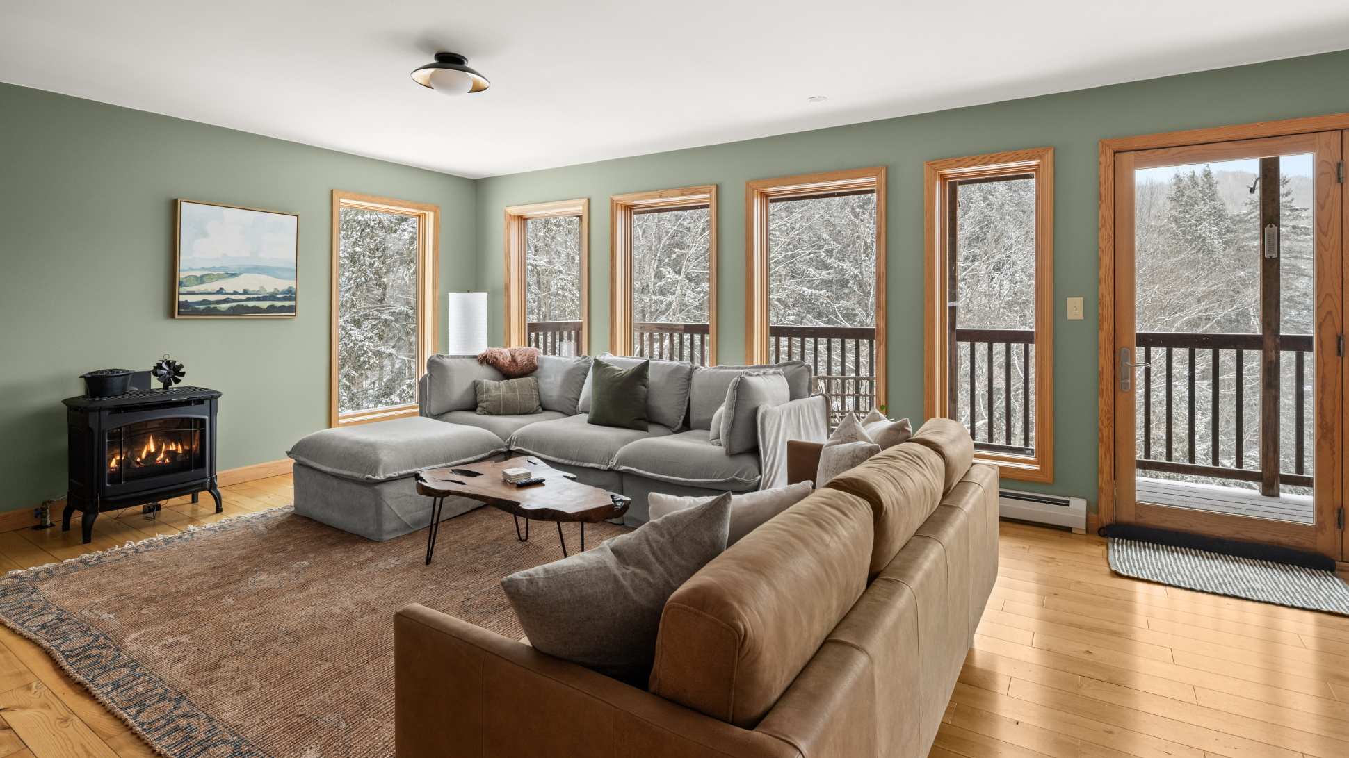 A spacious living room with light green walls, a large grey sectional sofa, a brown leather sofa, a unique live-edge coffee table, and a black wood-burning stove, all overlooking a snowy forest through multiple windows.