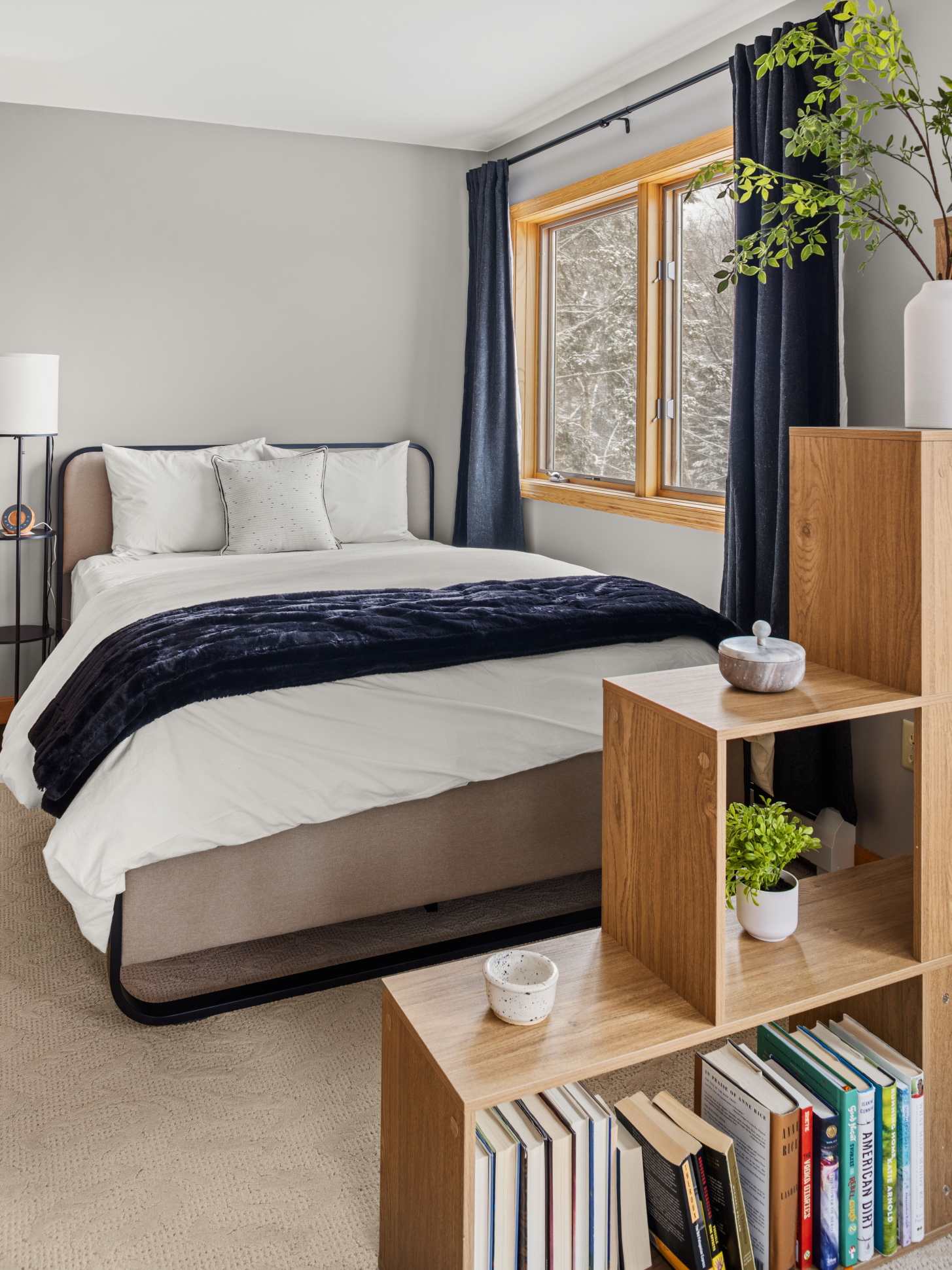 A serene bedroom features a queen-sized bed with white bedding and a dark blue throw, flanked by a modern black nightstand with a lamp. A large window with dark blue curtains provides a view of the snowy outdoors, and the room has light gray walls.