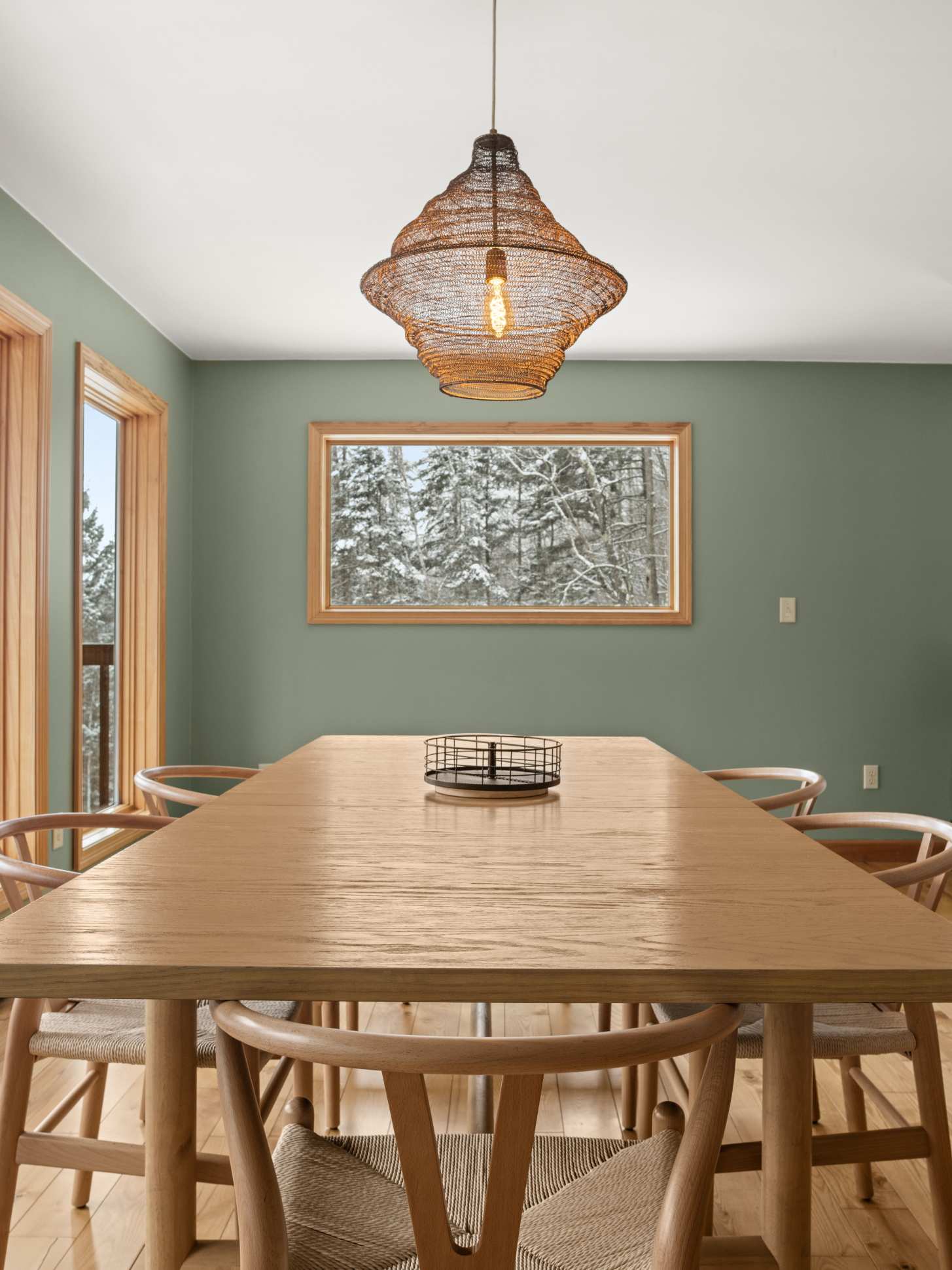 Close-up view of the wooden dining table with modern chairs and a woven pendant light, set against a light green wall with a framed window showcasing a snowy forest, emphasizing the natural light and rustic-modern aesthetic.