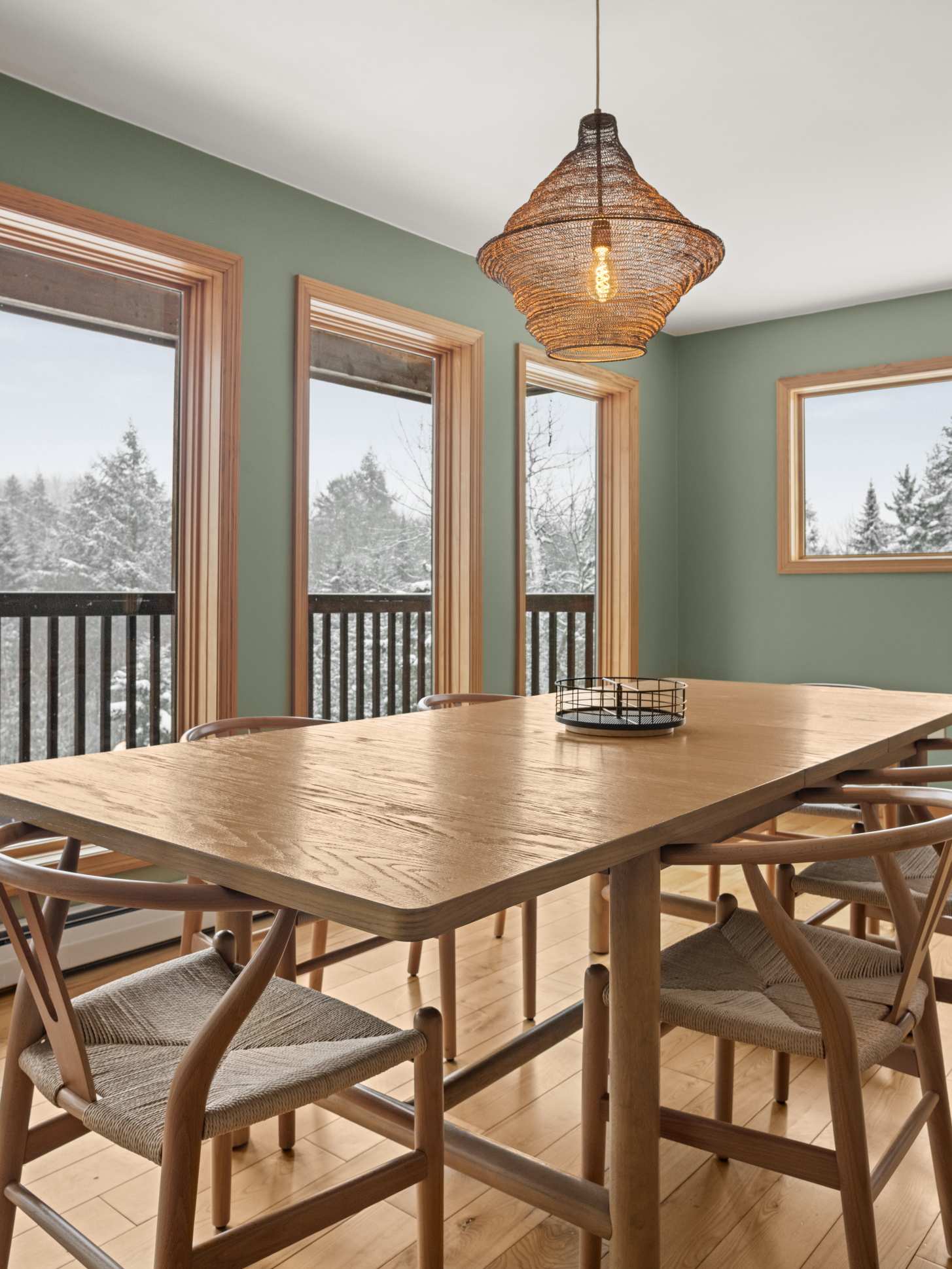 A close-up view of the dining area highlights the large light wood dining table and six matching chairs with woven seats. A unique woven pendant light illuminates the space, and large windows reveal a snowy forest outside.