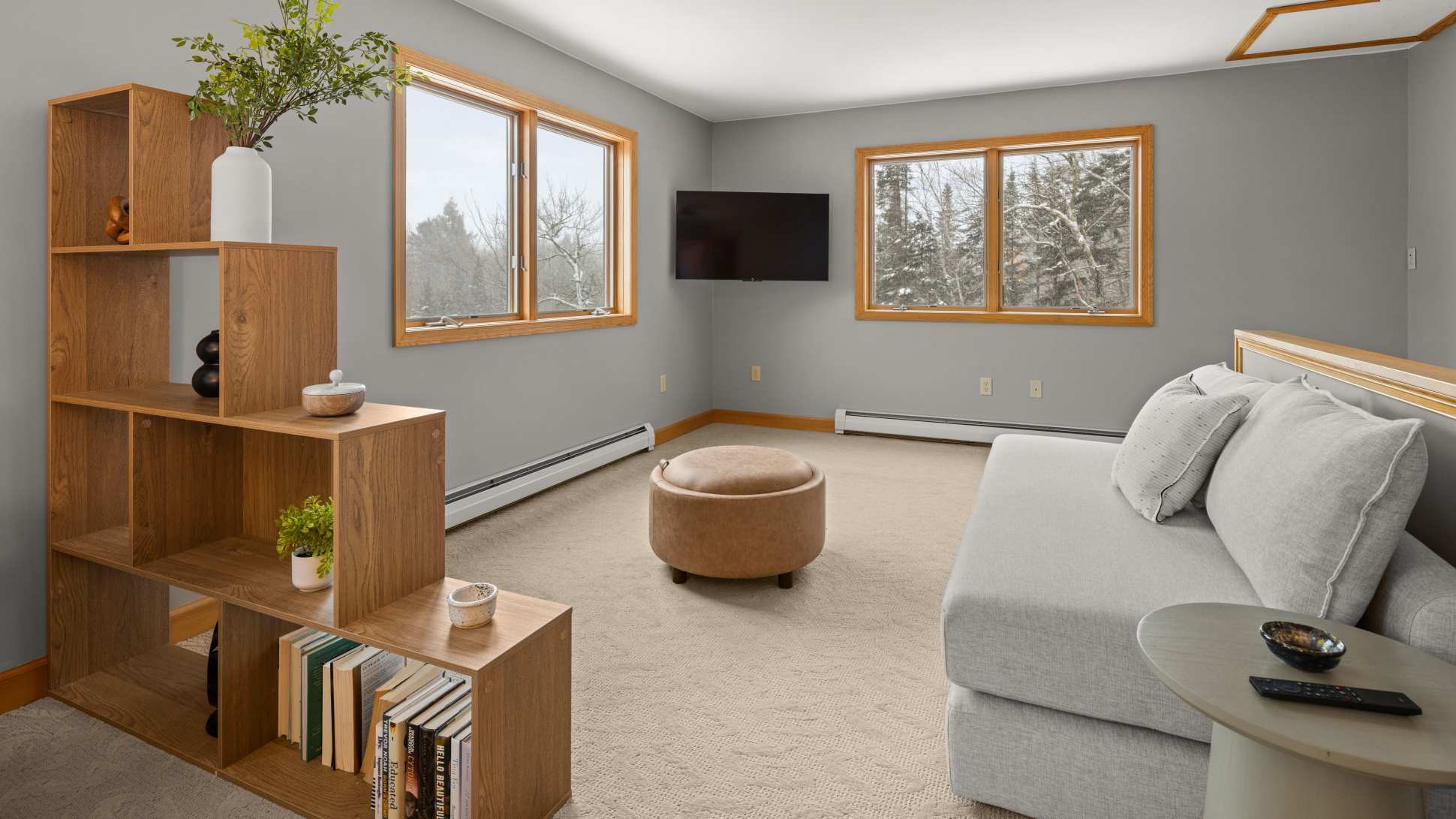 A bright living space with light grey walls and carpet, featuring a grey sectional sofa and a round leather ottoman. Large windows with natural wood trim offer views of snowy trees, and a wall-mounted TV is visible.