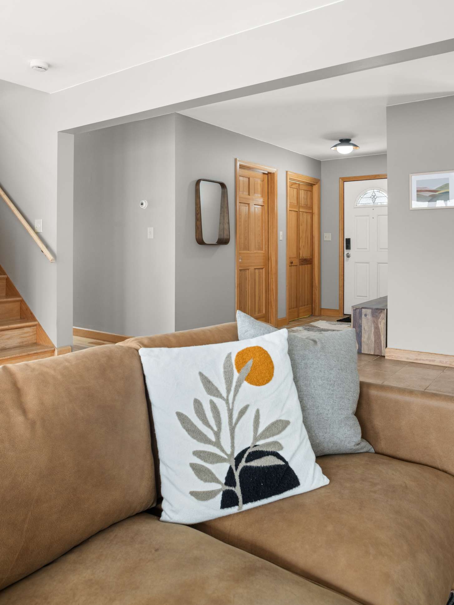 A view from the living room towards the entrance shows a light gray hallway with a wooden mirror and two wooden doors. The front door, partially visible, has a decorative glass insert, and the floor transitions from hardwood to tile.
