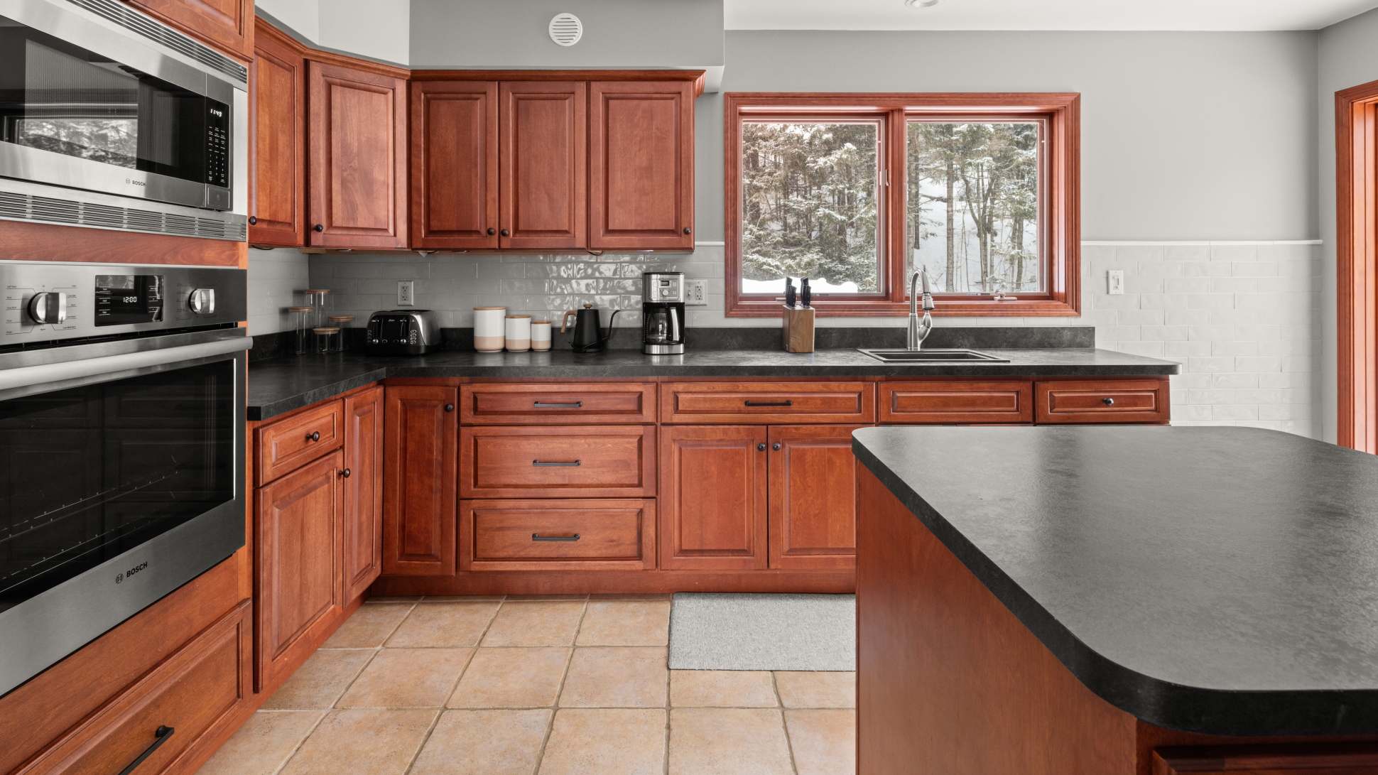 Traditional kitchen with warm cherry wood cabinetry, dark countertops, and a light gray subway tile backsplash, equipped with stainless steel Bosch appliances and a large window offering a view of a snowy outdoor scene.