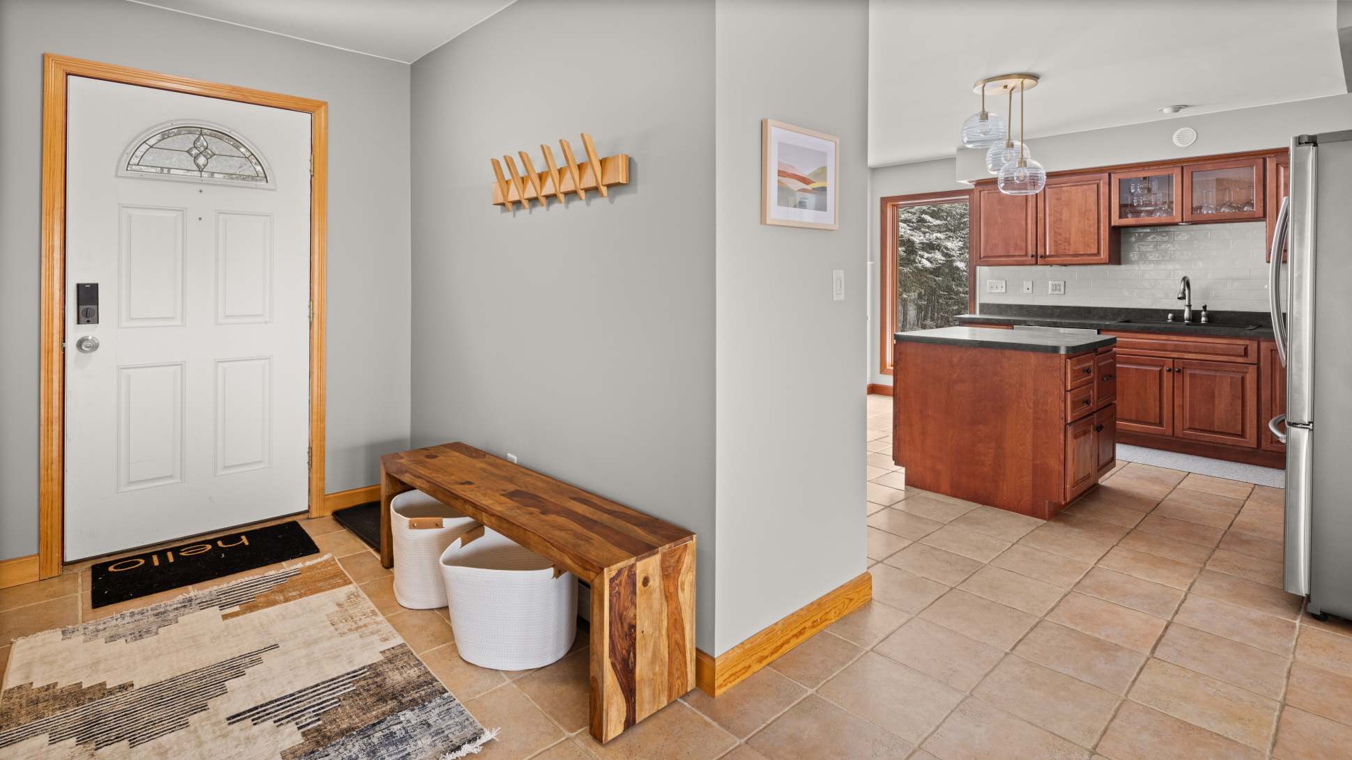 A welcoming foyer with light grey walls, a white front door with a decorative glass insert, a wooden bench, and a coat rack, leading into a kitchen with warm wood cabinetry.