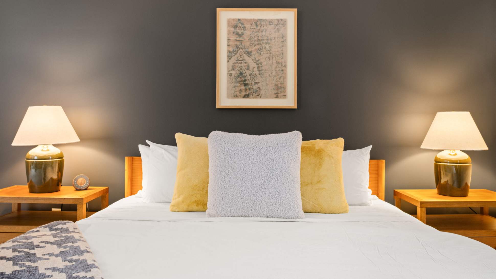 A cozy bedroom with a dark accent wall behind a queen-sized bed dressed in white linens and yellow and grey throw pillows. Two wooden nightstands with matching lamps flank the bed, beneath a framed artwork.