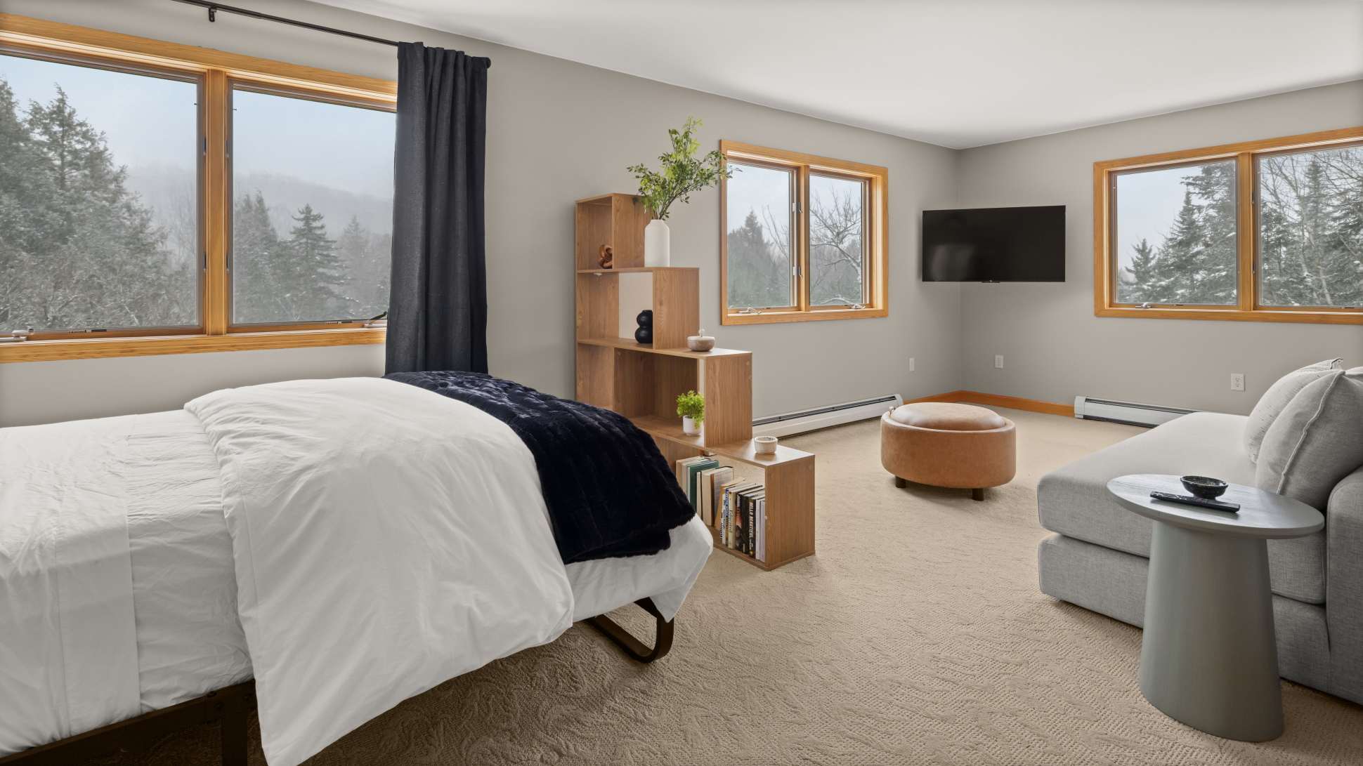 A spacious bedroom suite combining a sleeping area with a metal-framed bed and a sitting area with a grey sectional sofa and ottoman. Large windows with wood trim offer expansive views of snowy mountains and trees.
