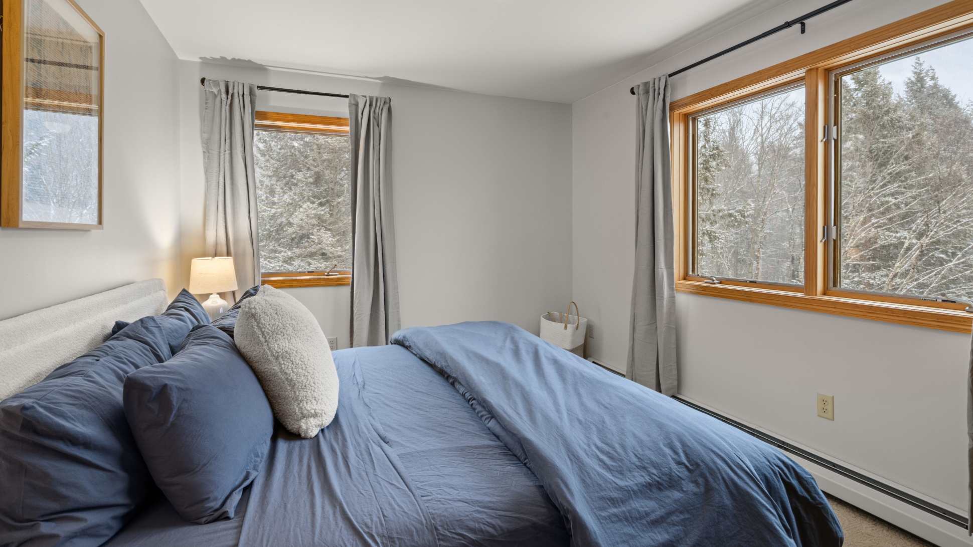 A cozy bedroom features a bed with blue bedding and a white textured pillow, positioned between two large windows with light gray curtains. The windows offer a view of a snowy forest, and the room has light gray walls and hardwood floors.