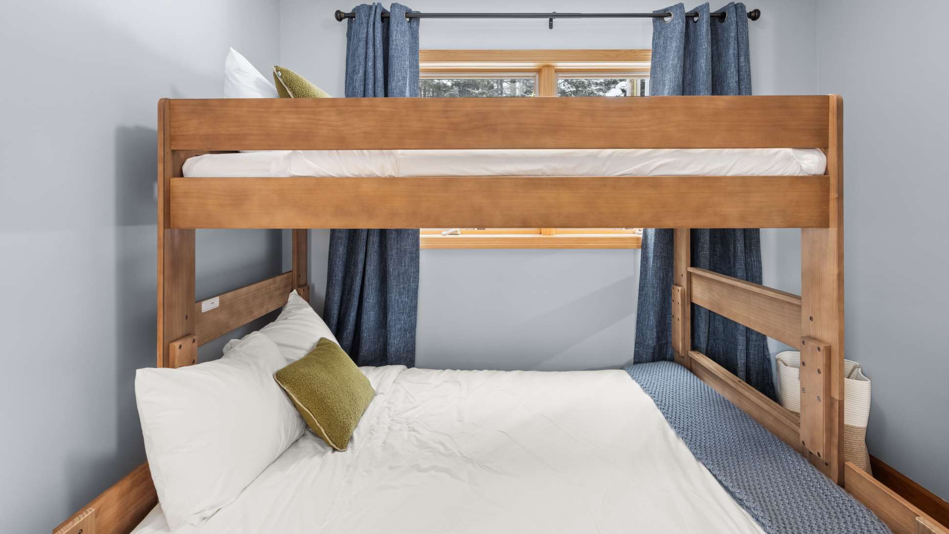 A bedroom designed for multiple guests features a sturdy wooden bunk bed with white bedding and a blue throw blanket on the lower bunk. Blue curtains frame a window offering a view of the snowy outdoors.