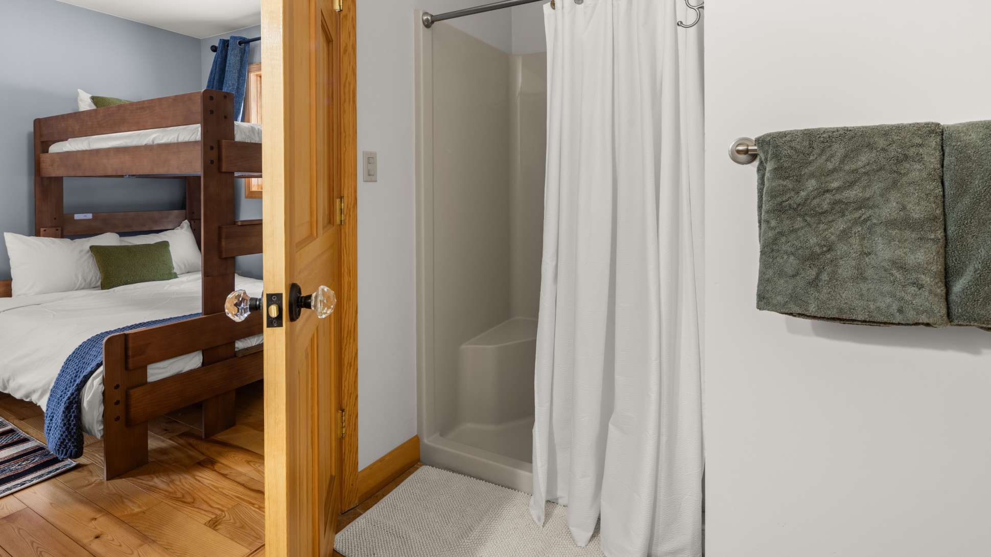 A simple bathroom features a white shower stall with a white curtain, and a white bath mat on the floor. A wooden door with a crystal knob is open, revealing a glimpse of a bedroom with bunk beds.
