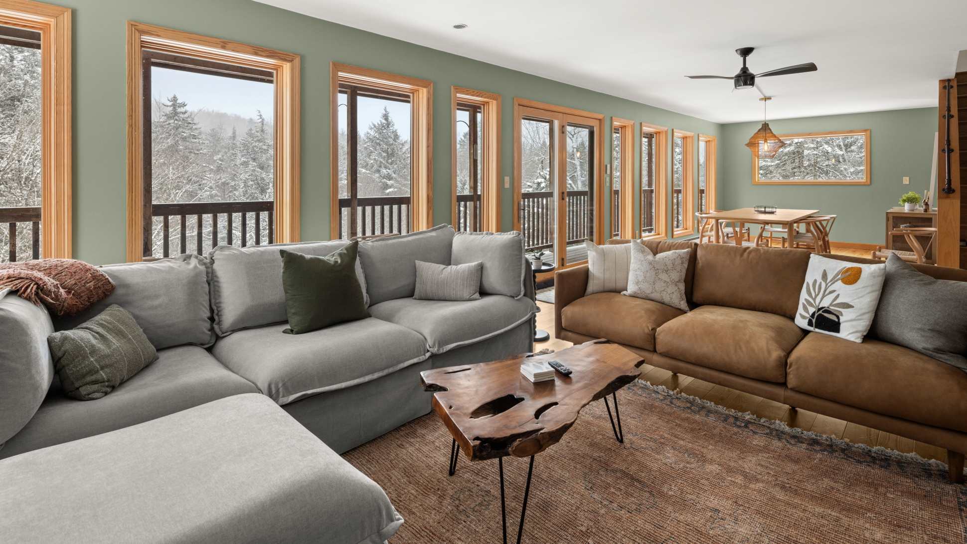 An expansive living room with light green walls, a large grey sectional and brown leather sofa, a live-edge coffee table, and multiple windows and a glass door leading to a balcony, offering snowy forest views.