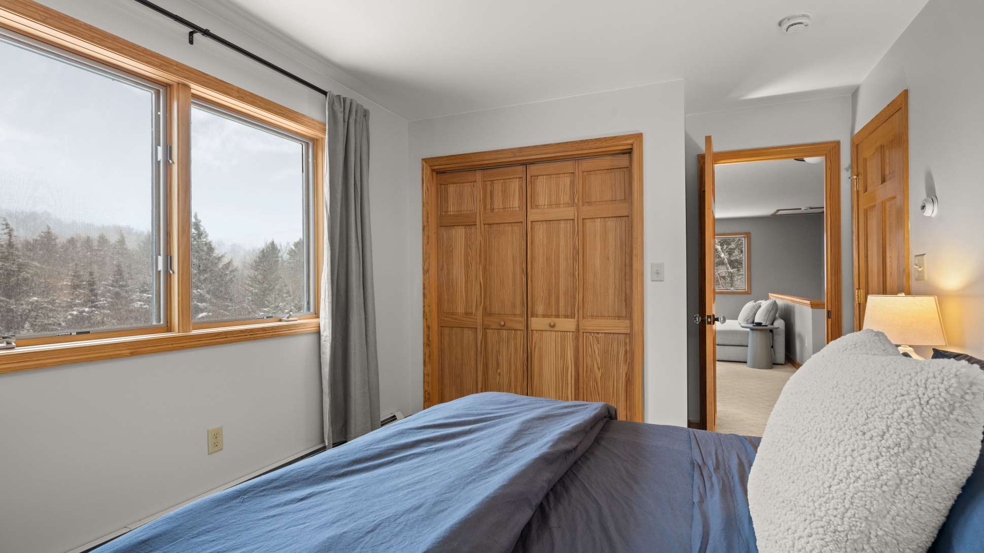 A cozy bedroom with a bed dressed in blue, flanked by wooden nightstands. A large window with grey curtains offers a view of snowy trees, and a wooden bi-fold closet door is visible.