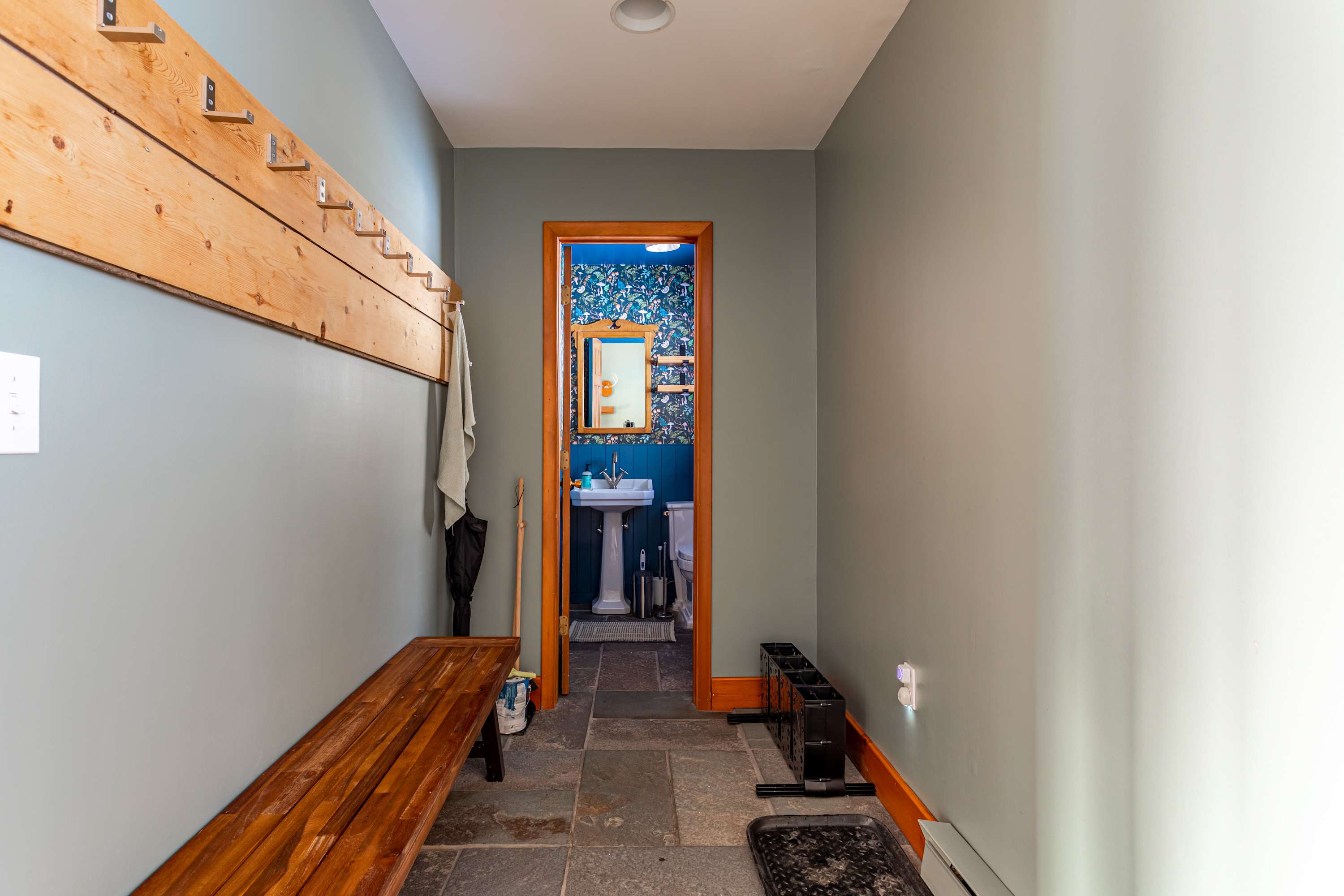 A practical mudroom with slate floor tiles, a long wooden bench, and coat hooks along one wall. An open doorway provides a view into a distinct powder room with mushroom wallpaper and a pedestal sink.