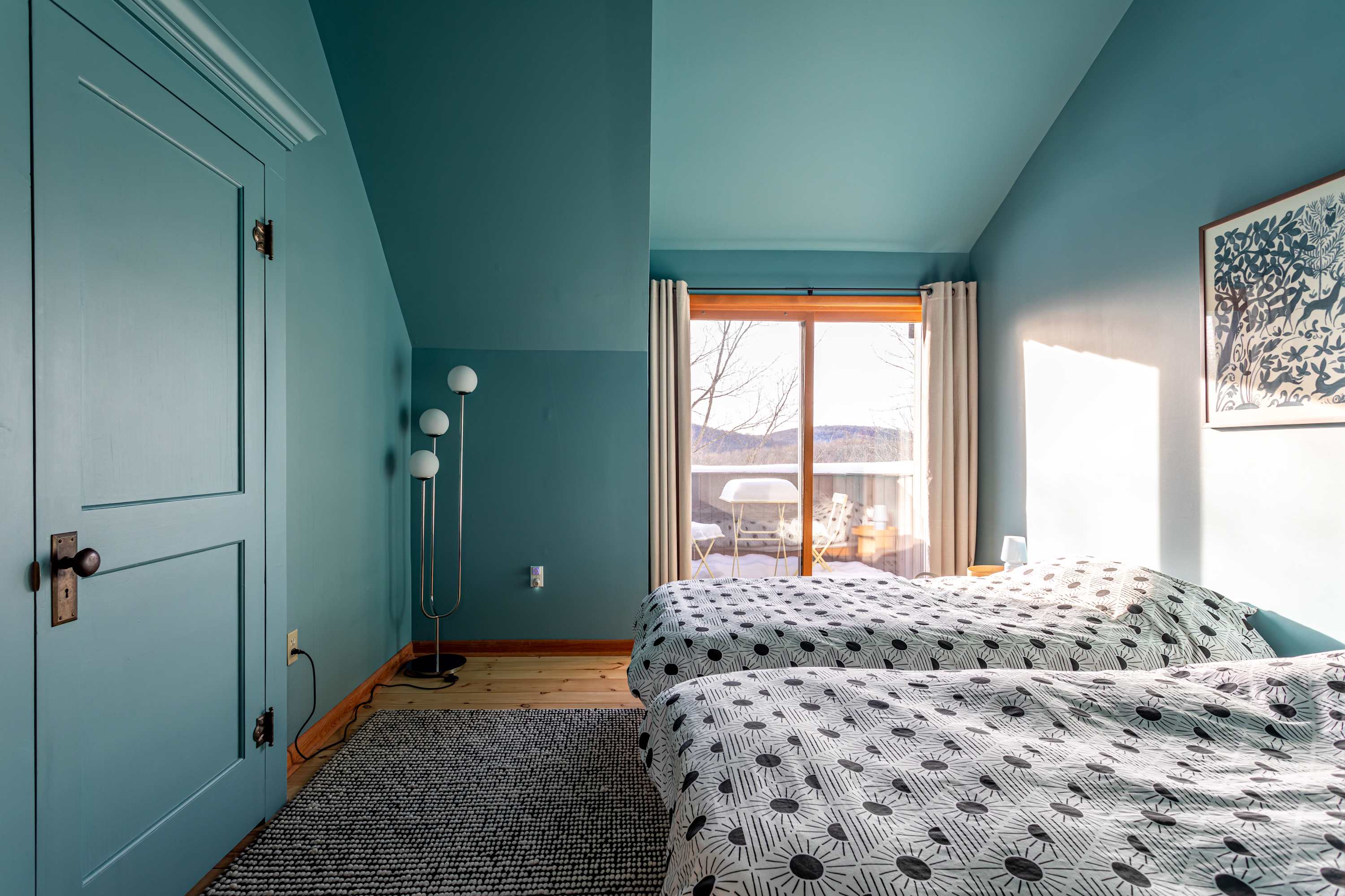 A serene bedroom with calming blue walls and ceiling, featuring two single beds with modern patterned bedding. A sliding glass door opens to a private balcony with outdoor seating and scenic views.