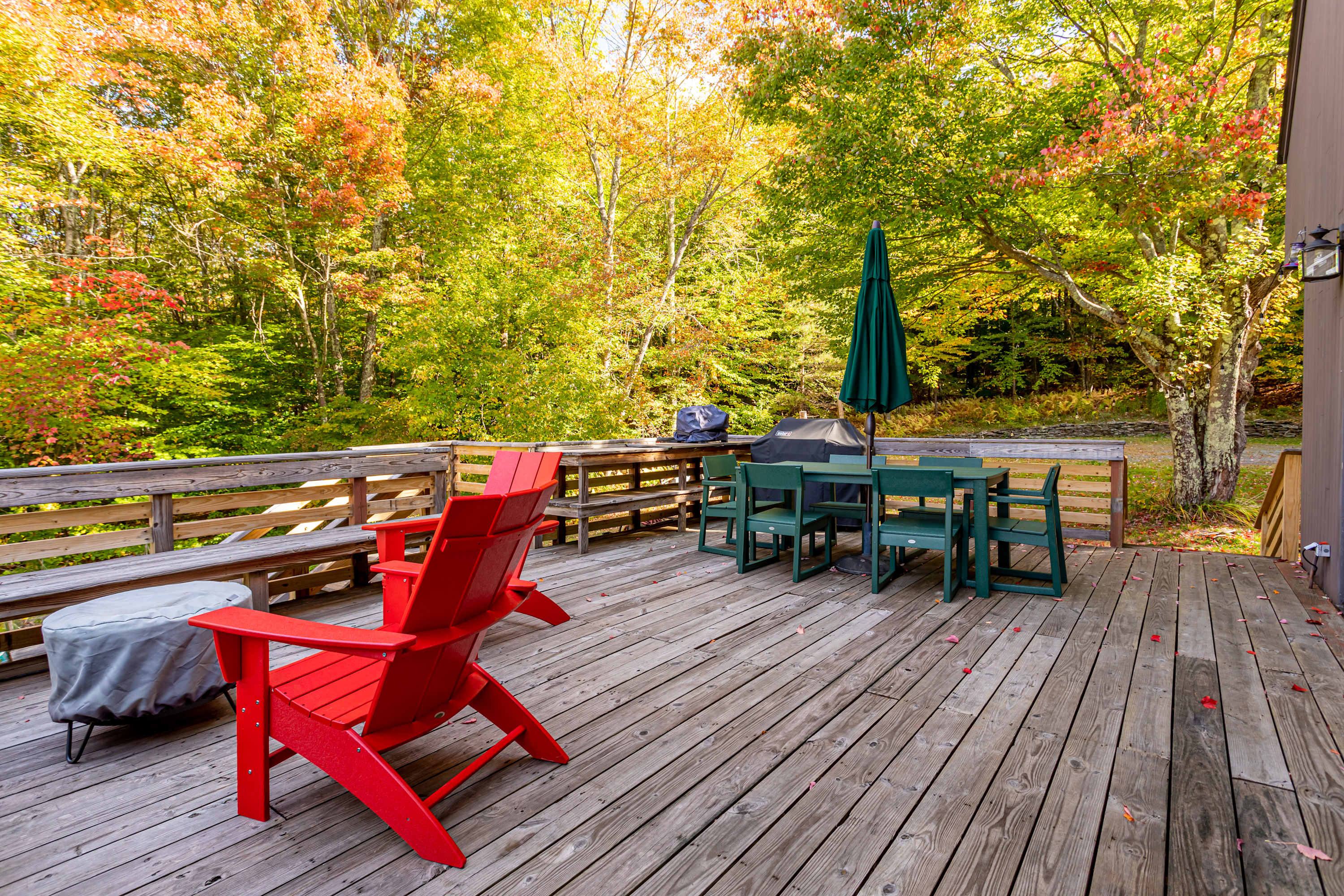 A spacious wooden deck features a dining area with a green table and chairs, a BBQ grill, and two bright red Adirondack chairs next to a covered fire pit, all surrounded by colorful autumn trees.