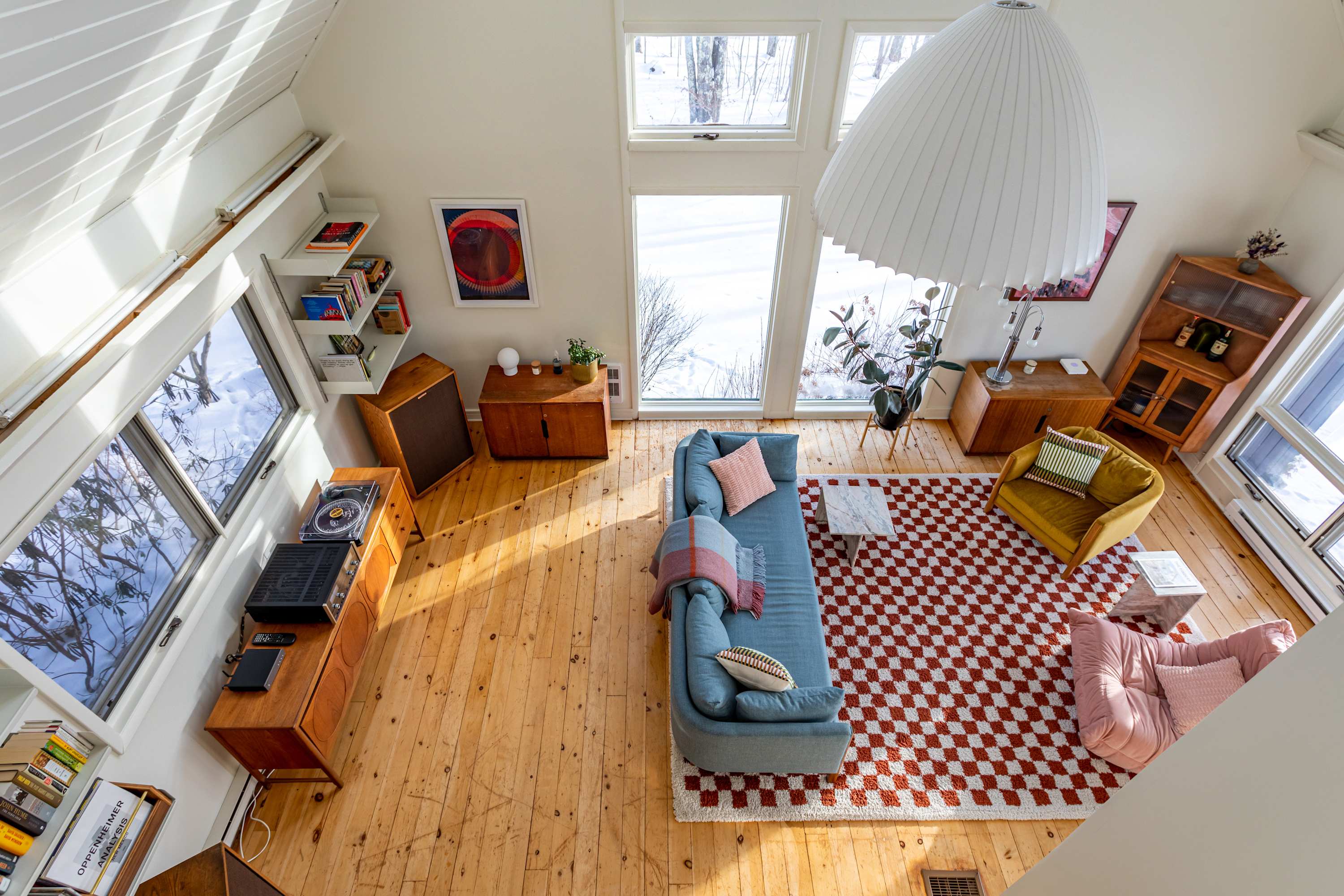 A bright, high-ceilinged living room with light hardwood floors, a blue sofa, and a red and white checkered rug. Large windows offer views of a snowy landscape, and a distinctive white pendant light hangs from above.