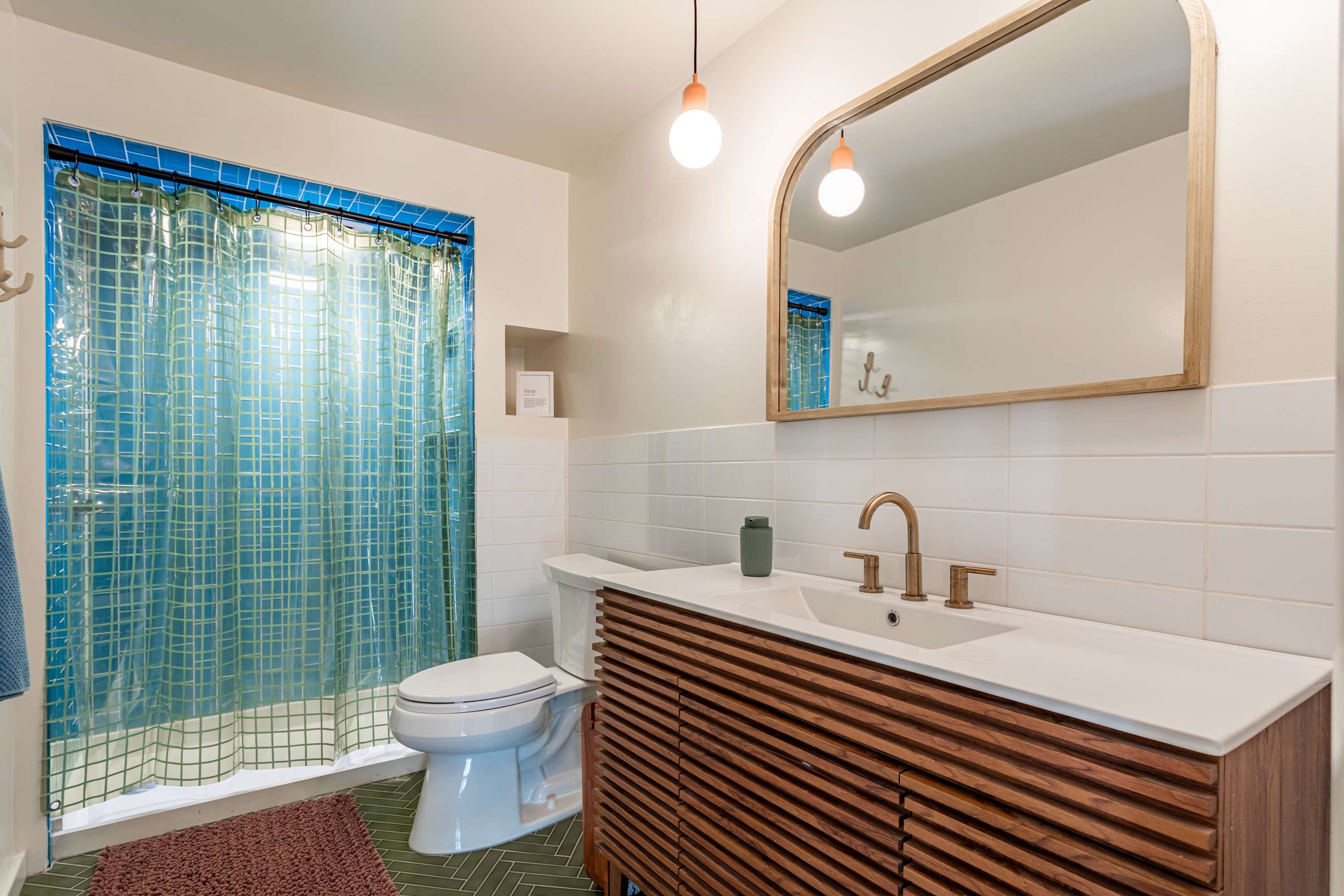A modern bathroom boasts a slatted wood vanity with a white countertop and a large arched mirror. The shower area is tiled in blue subway tiles with a translucent blue grid curtain, and the floor features green hexagonal tiles.