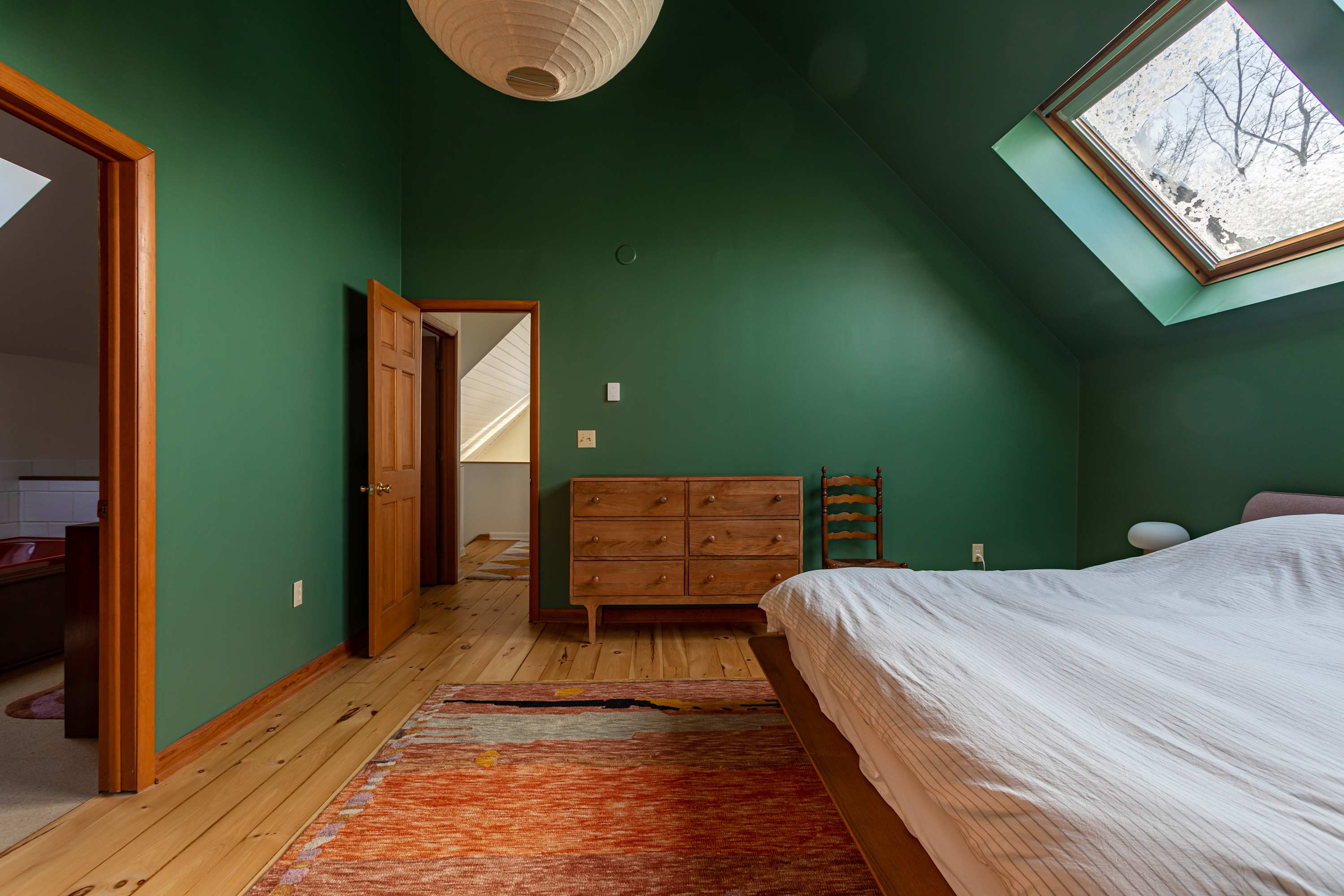 Another view of the dark green bedroom, showcasing a vintage wooden dresser with multiple drawers and a small wooden chair. A skylight and a paper lantern light fixture illuminate the space, with an area rug on the wood floor.