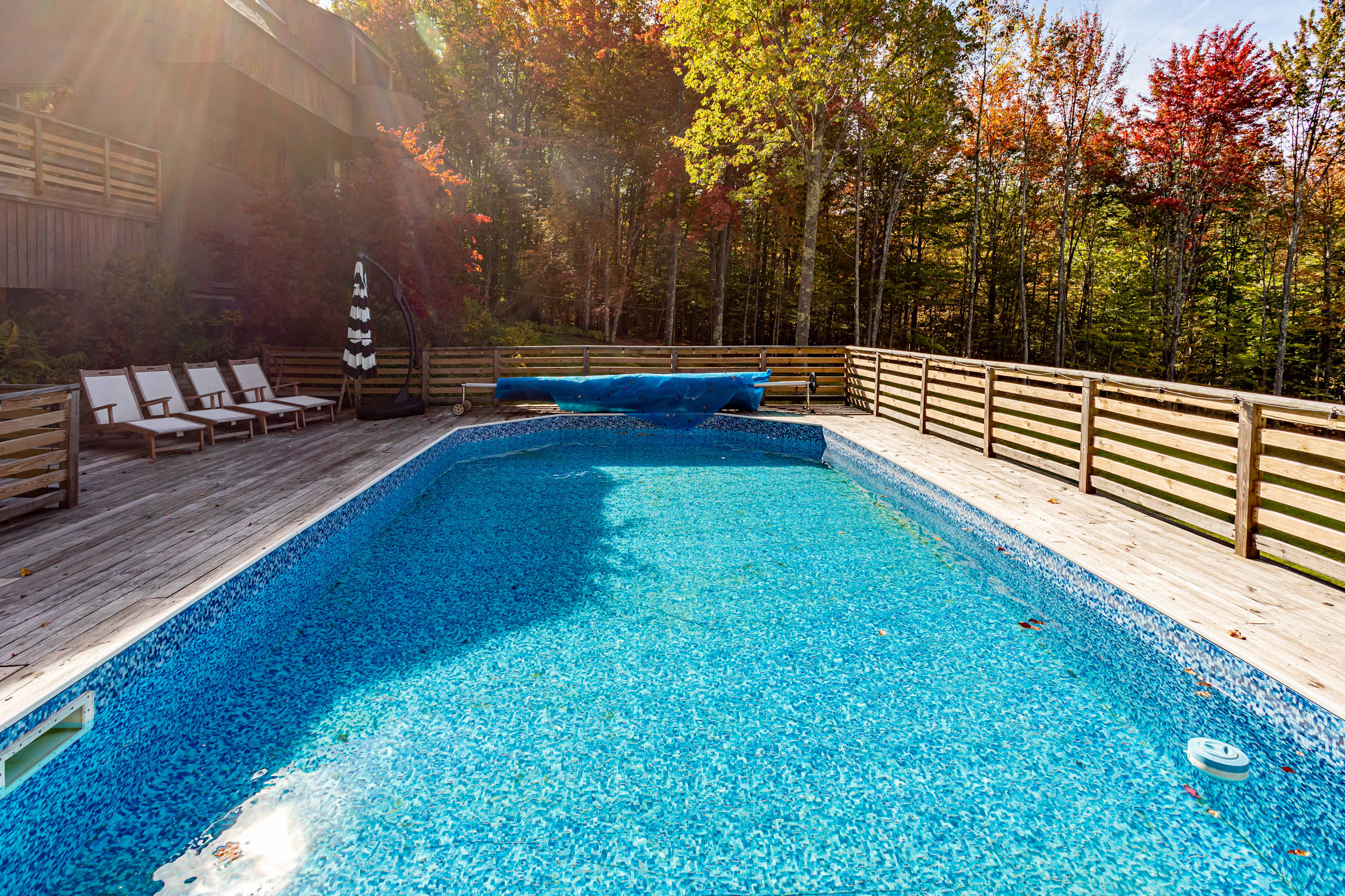 An inviting rectangular swimming pool with a blue liner is surrounded by a spacious wooden deck, featuring white lounge chairs and an umbrella, set against a vibrant backdrop of autumn trees.