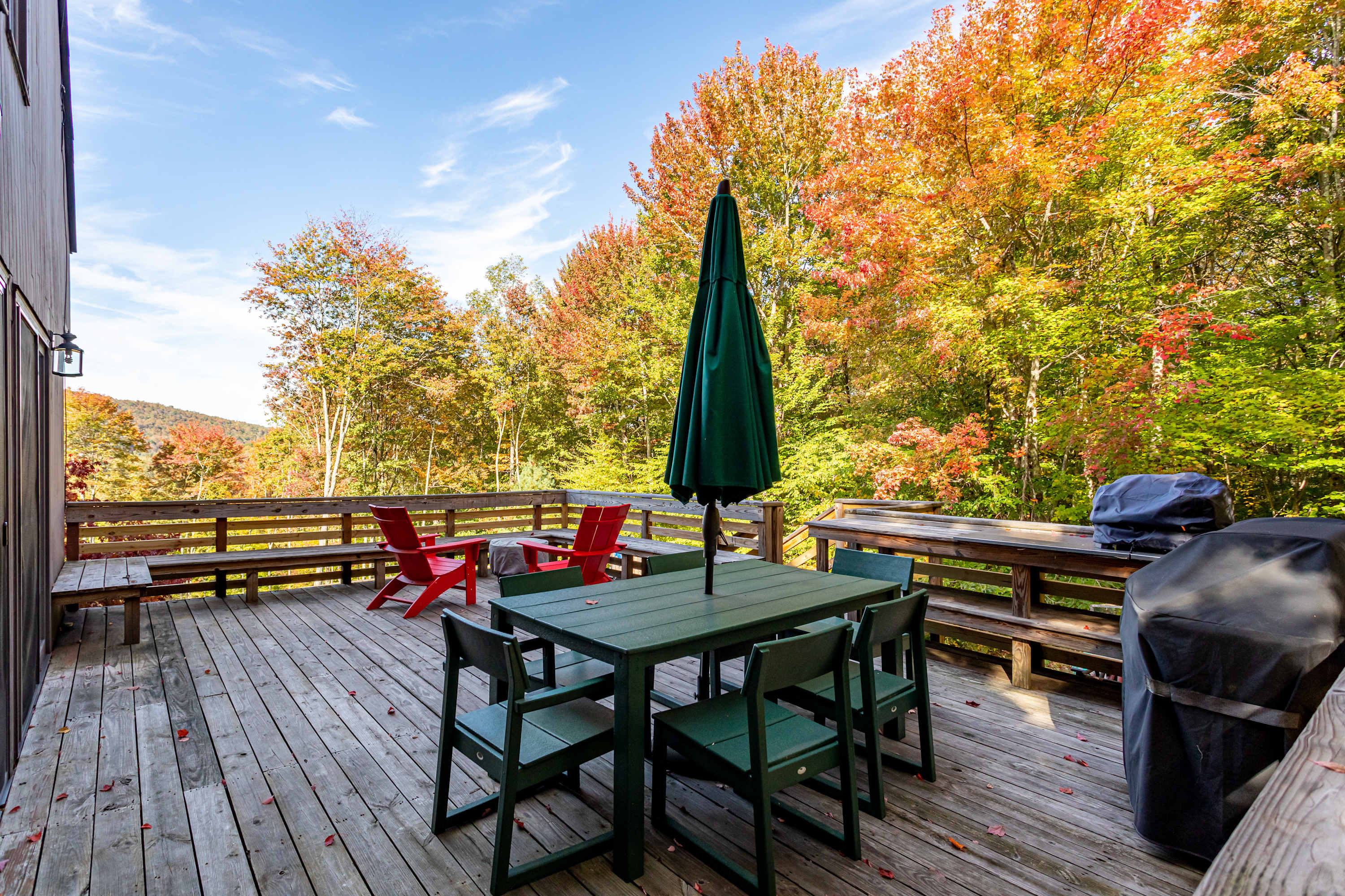 A spacious wooden deck with a green outdoor dining set, red Adirondack chairs, and a grill, is surrounded by vibrant autumn trees and offers scenic views of rolling hills.