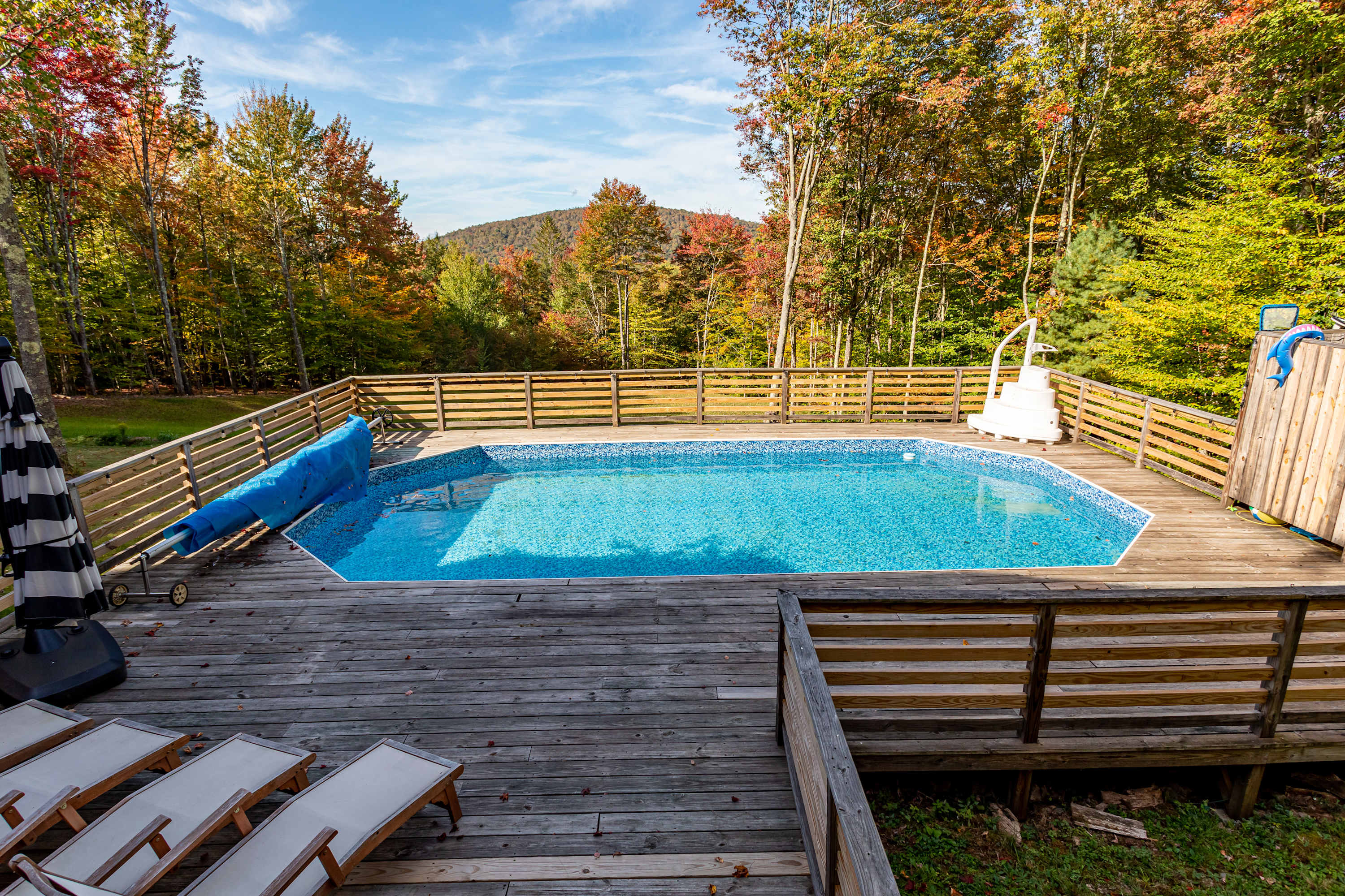 An elevated view of a blue swimming pool surrounded by a wooden deck and fence, with a distant mountain range visible beyond the colorful autumn trees. A white pool ladder and a blue pool cover are also present.