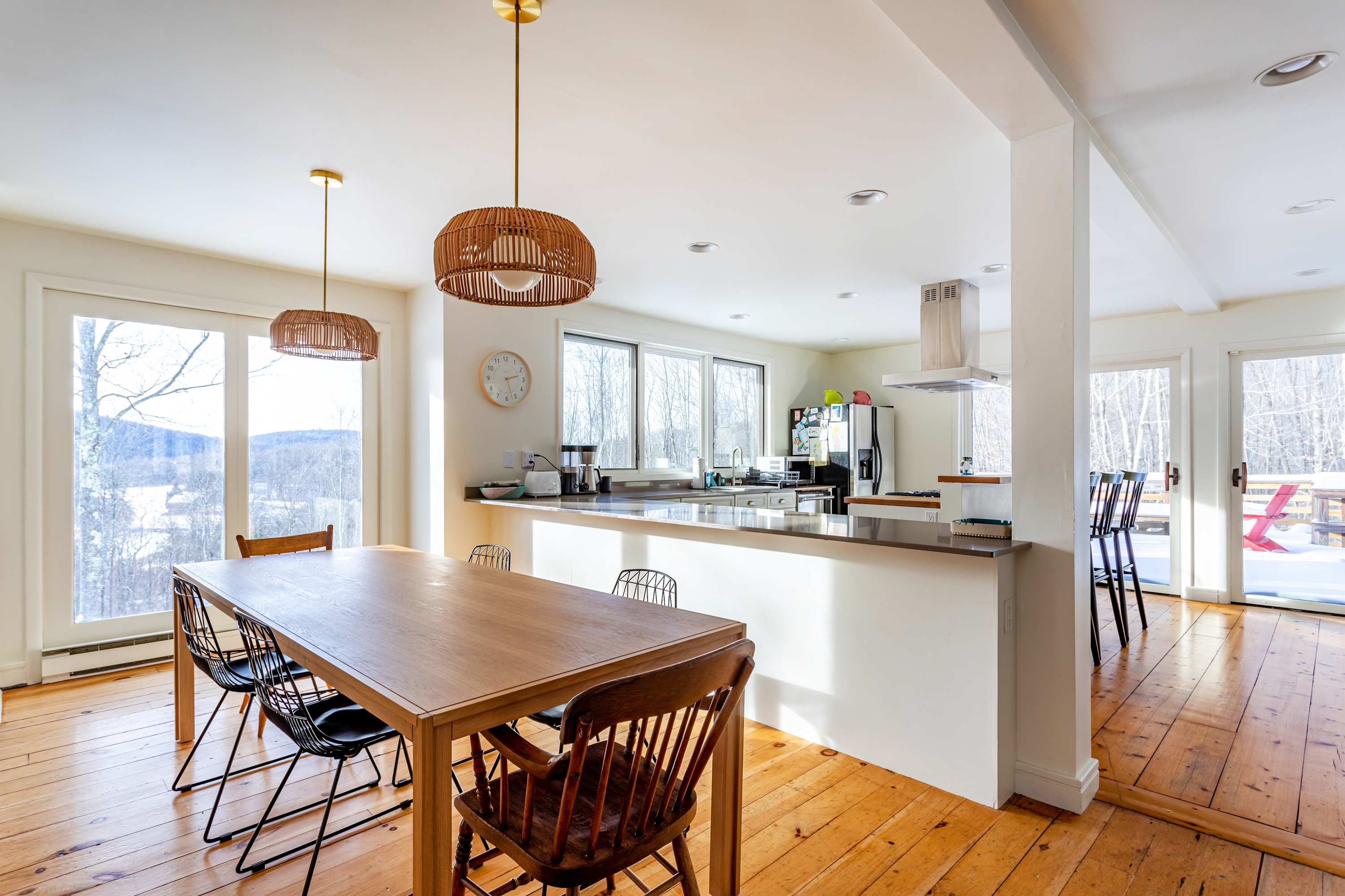 A bright dining area with a long wooden table and mixed chairs, illuminated by two woven pendant lights. It opens into a modern kitchen with white cabinetry and large windows overlooking a snowy landscape.