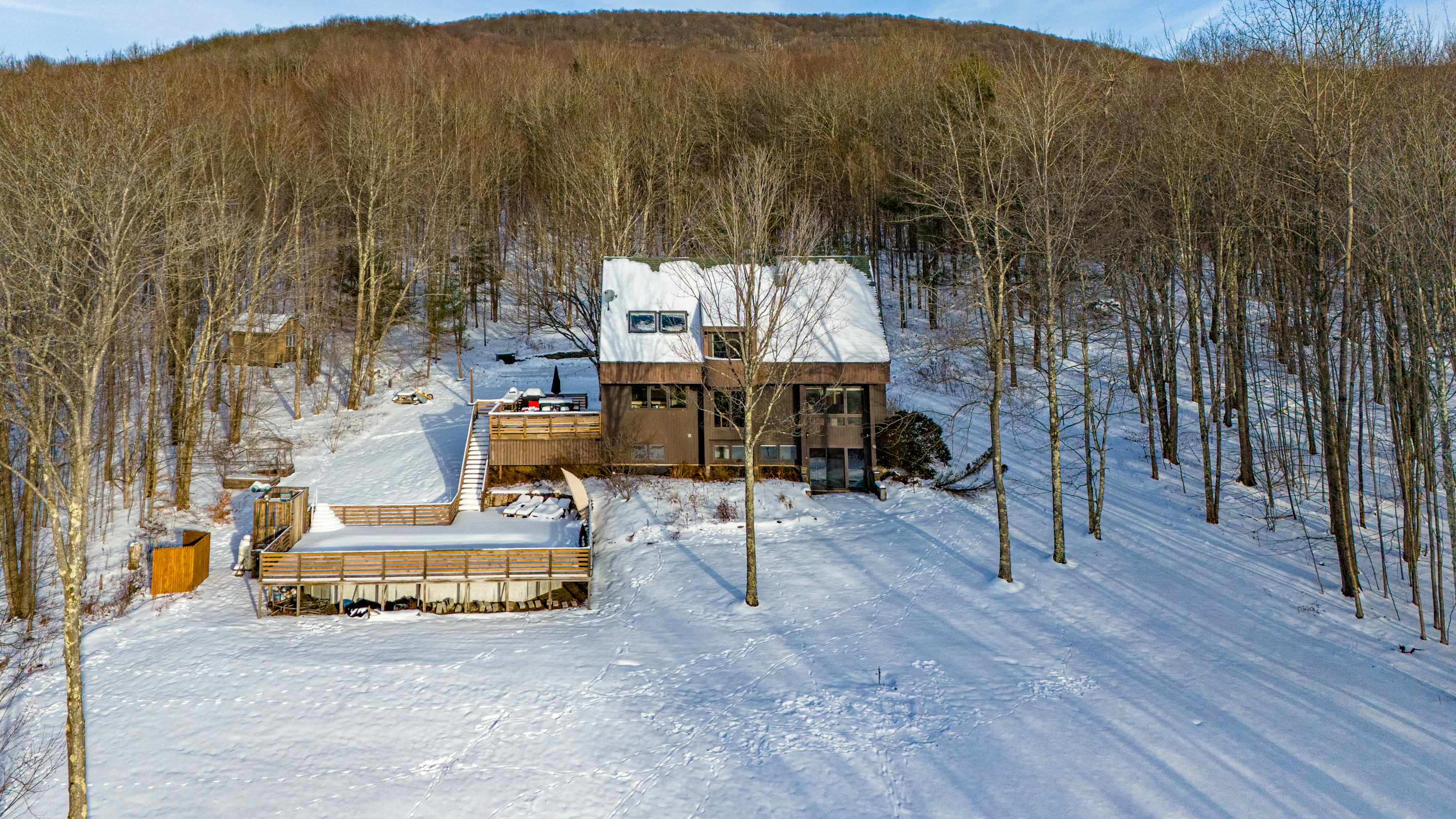 An aerial perspective reveals a brown A-frame house nestled on a snow-covered hillside, surrounded by a dense forest of bare trees. A large, multi-level wooden deck extends from the property, offering expansive views of the winter landscape.