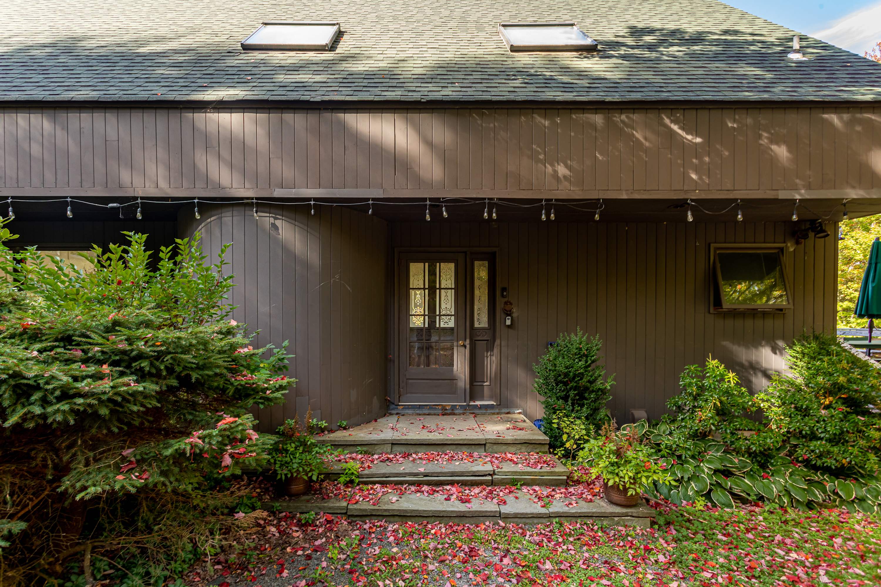 A close-up of the front entrance of the brown house, featuring a dark wood door with decorative glass panels and two skylights on the dark green roof. The entrance is adorned with string lights, potted plants, and surrounded by green bushes and red autumn leaves on the ground.