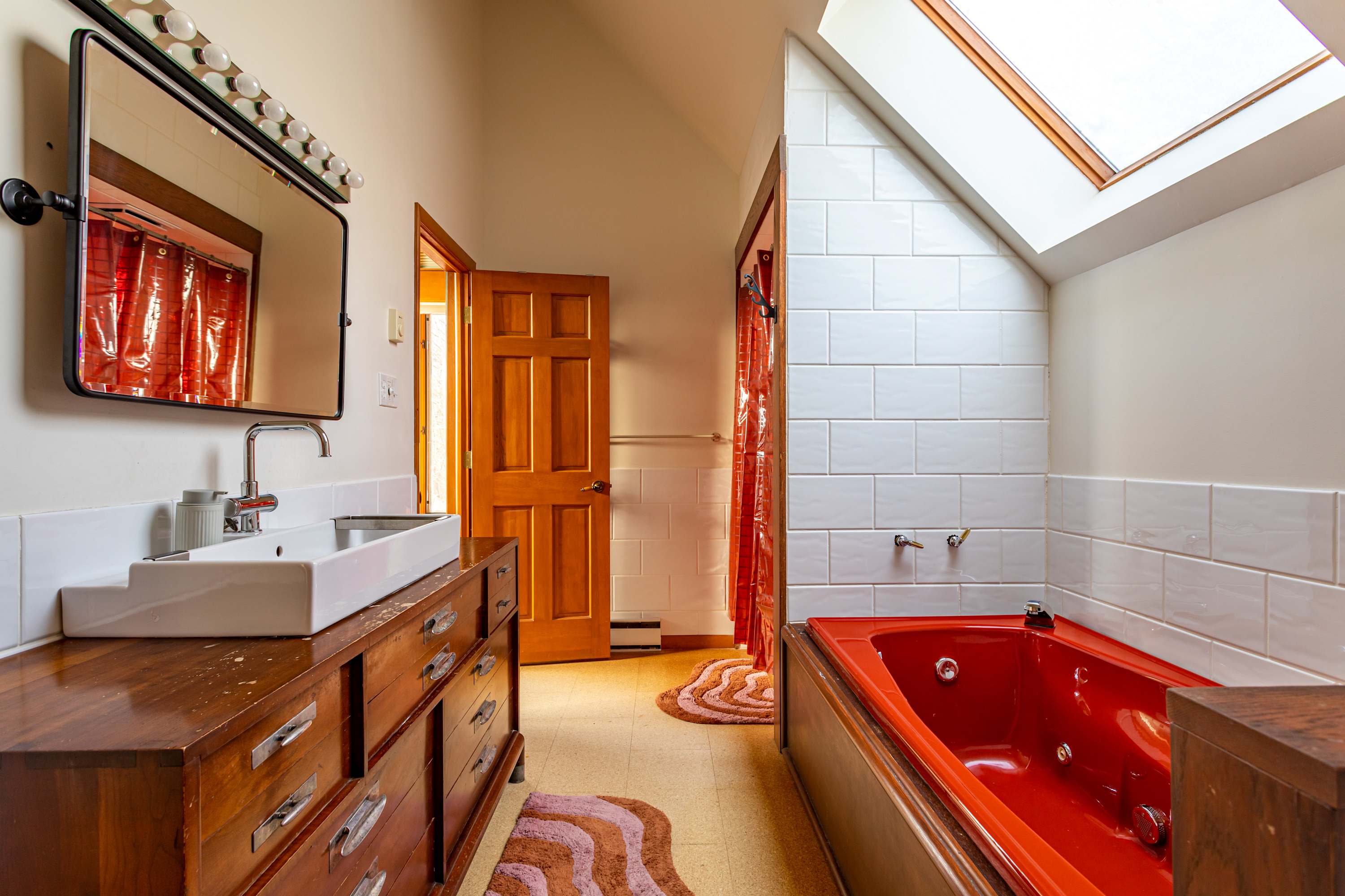 Another angle of the bathroom, highlighting the red jetted tub with a matching red shower curtain. The wooden vanity with a white sink and mirror is visible, alongside white tiled walls and a skylight.