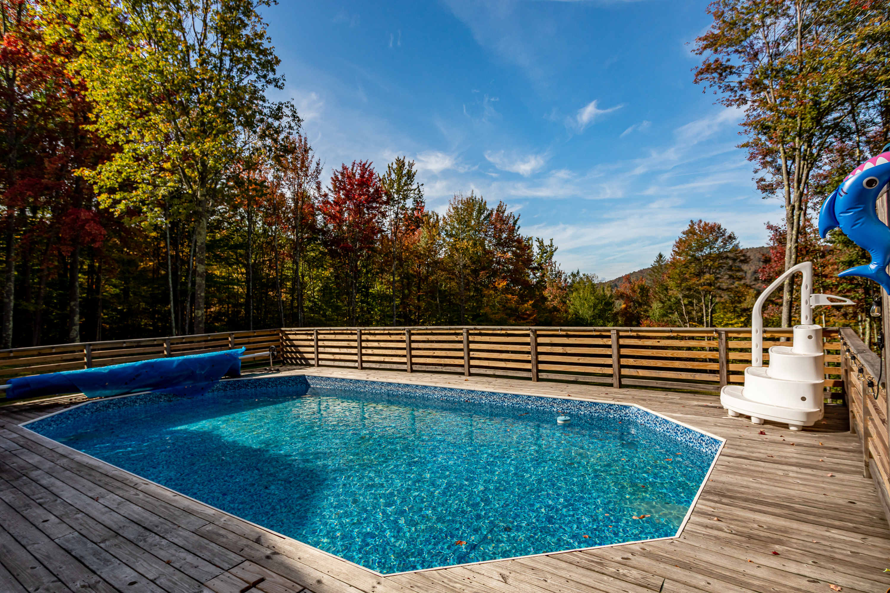 A bright blue swimming pool is set within a wooden deck, enclosed by a slatted wooden fence, with colorful autumn trees and a clear blue sky in the background. A white pool ladder and a blue inflatable dolphin are visible.